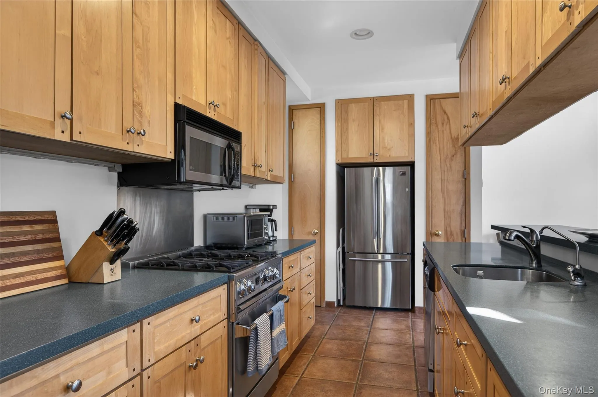 Galley kitchen featuring natural wood cabinetry, dark countertops, stainless steel appliances, and tiled flooring Galley kitchen featuring natural wood cabinetry, dark countertops, stainless steel appliances, and tiled flooring