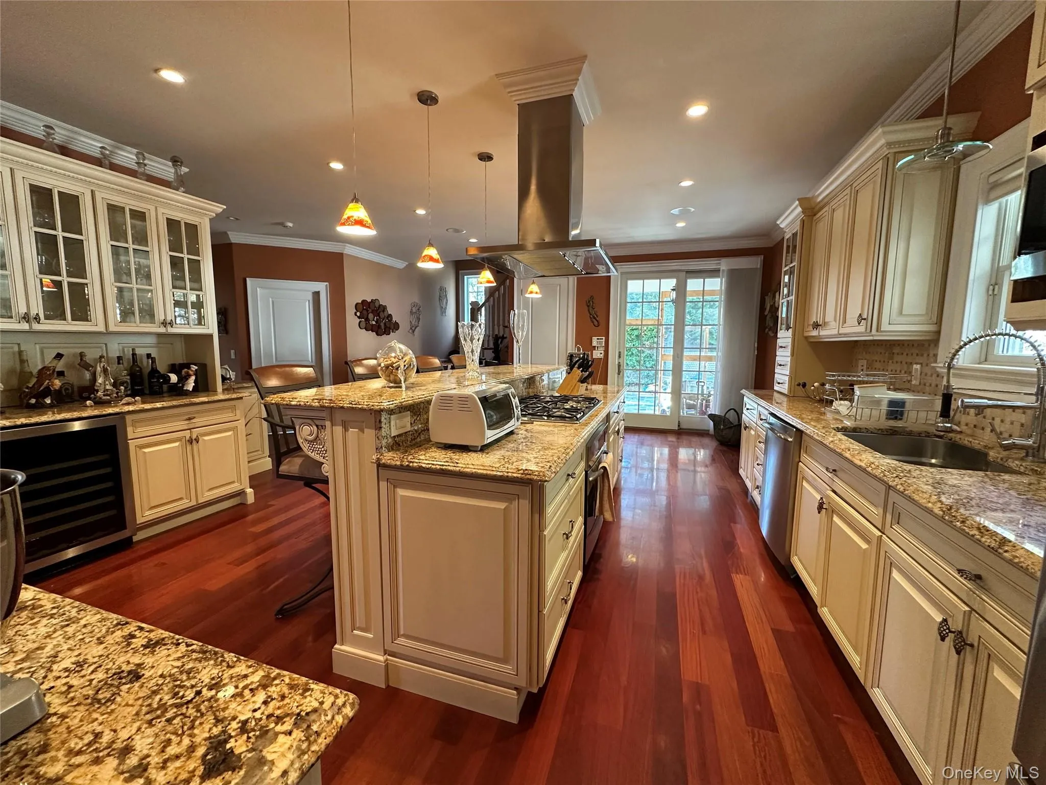 Kitchen with custom cabinetry, light granite countertops, Turkish marble mosaic backsplash, crown molding, and island range hood, ornamental molding, light granite counters, beverage cooler, and pendant lighting Kitchen with custom cabinetry, light granite countertops, Turkish marble mosaic backsplash, crown molding, and island range hood, ornamental molding, light granite counters, beverage cooler, and pendant lighting