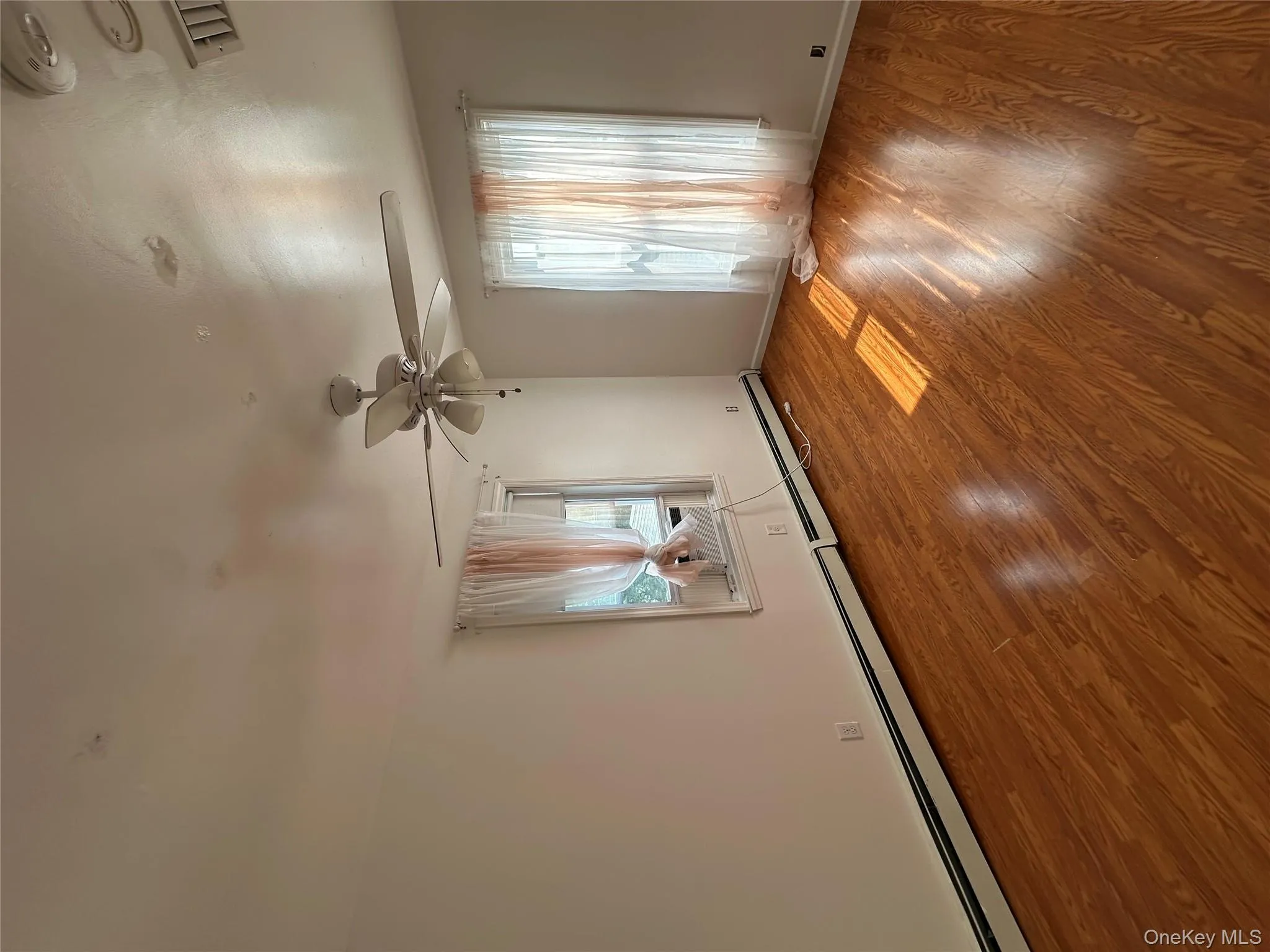 Unfurnished room with healthy amount of natural light, wood finished floors, a ceiling fan, and baseboard heating Unfurnished room with healthy amount of natural light, wood finished floors, a ceiling fan, and baseboard heating