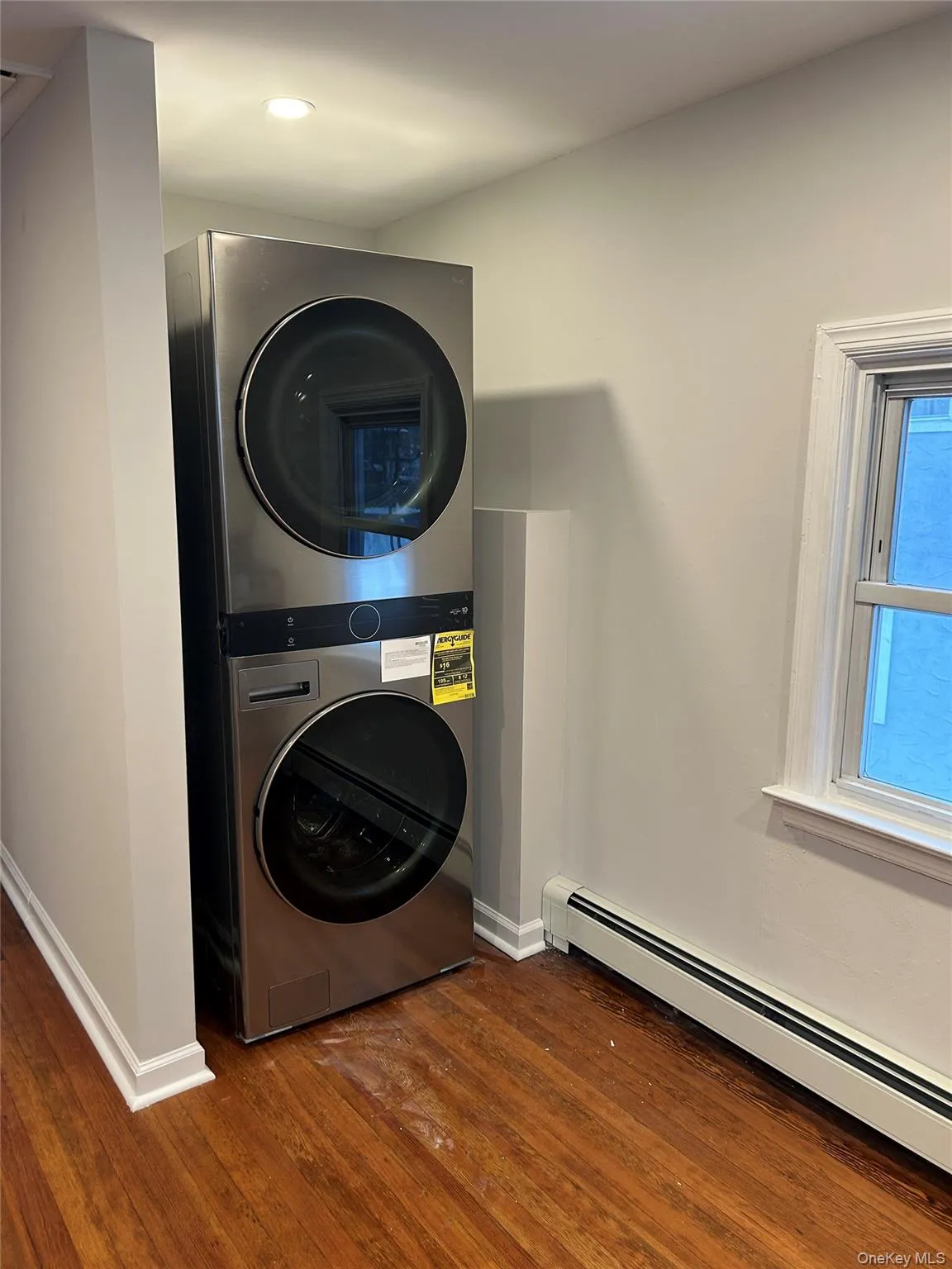Washroom featuring a baseboard heating unit, stacked washer and clothes dryer, and dark wood-style flooring Washroom featuring a baseboard heating unit, stacked washer and clothes dryer, and dark wood-style flooring