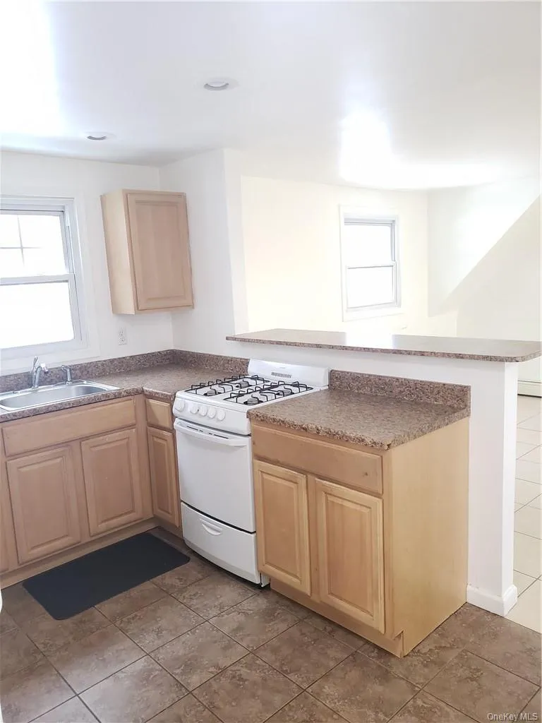 Kitchen with light wood finish cabinets, white range with gas cooktop, a peninsula, dark countertops, and light tile patterned flooring Kitchen with light wood finish cabinets, white range with gas cooktop, a peninsula, dark countertops, and light tile patterned flooring