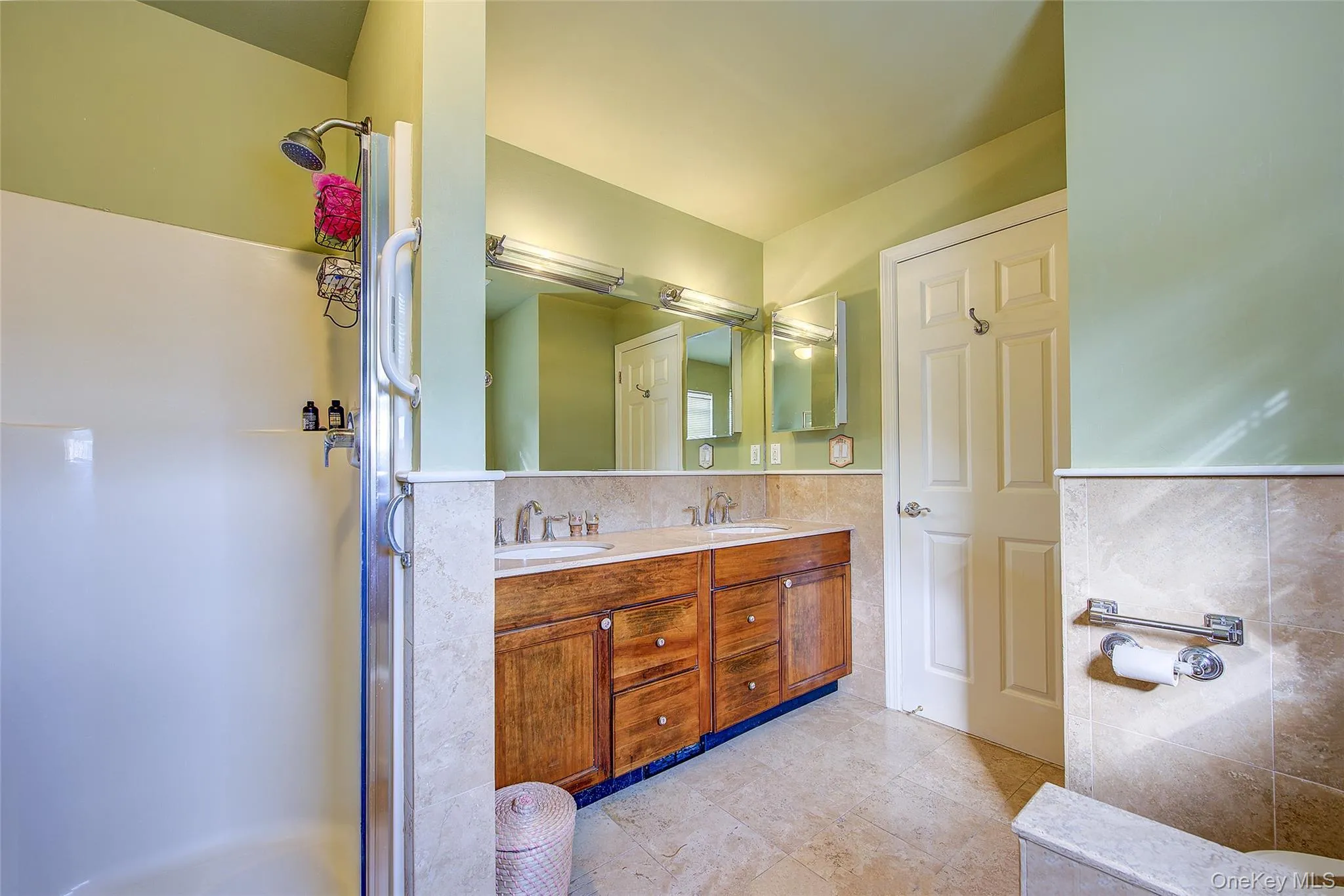 Bathroom featuring tile walls, double vanity, a shower stall, a wainscoted wall, and light tile patterned floors Bathroom featuring tile walls, double vanity, a shower stall, a wainscoted wall, and light tile patterned floors