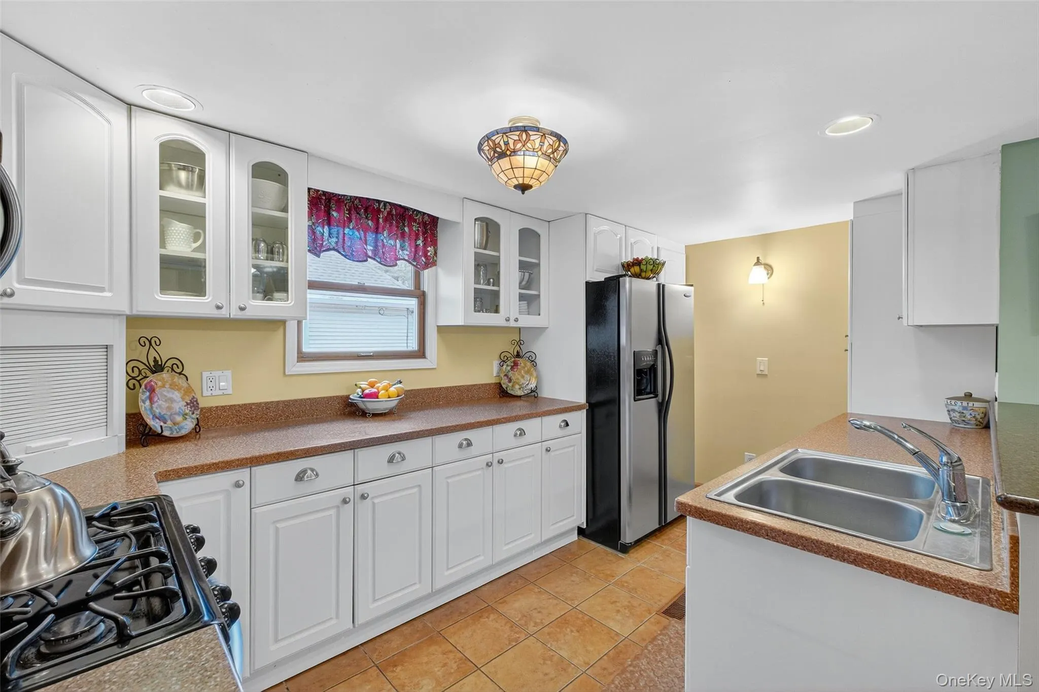 Kitchen with white cabinetry, stainless steel fridge with ice dispenser, glass insert cabinets, black gas range oven, and light tile patterned floors Kitchen with white cabinetry, stainless steel fridge with ice dispenser, glass insert cabinets, black gas range oven, and light tile patterned floors