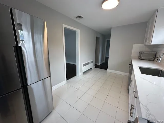 Kitchen featuring freestanding refrigerator, white cabinetry, light tile patterned flooring, light stone counters, and radiator heating unit Kitchen featuring freestanding refrigerator, white cabinetry, light tile patterned flooring, light stone counters, and radiator heating unit