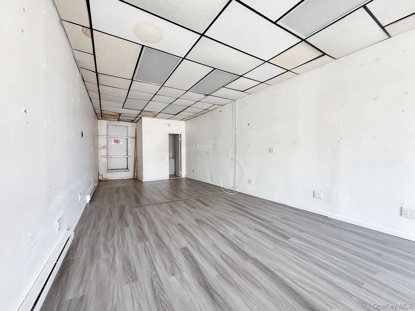 Empty commercial space with baseboard heating, light wood-style flooring, and a drop ceiling Empty commercial space with baseboard heating, light wood-style flooring, and a drop ceiling