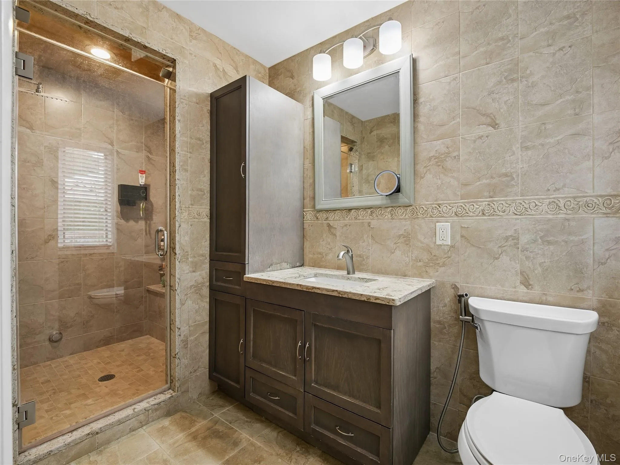 Bathroom featuring floor-to-ceiling tile, a frameless glass shower enclosure, a vanity with a light-toned countertop, and a built-in storage cabinet and steam shower. Bathroom featuring floor-to-ceiling tile, a frameless glass shower enclosure, a vanity with a light-toned countertop, and a built-in storage cabinet and steam shower.