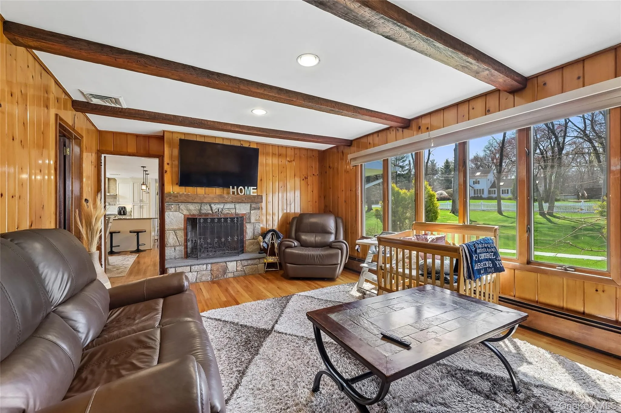 Living area featuring wood-finish wall paneling, a stone fireplace, exposed ceiling beams, hardwood-finish flooring, and expansive windows. Living area featuring wood-finish wall paneling, a stone fireplace, exposed ceiling beams, hardwood-finish flooring, and expansive windows.