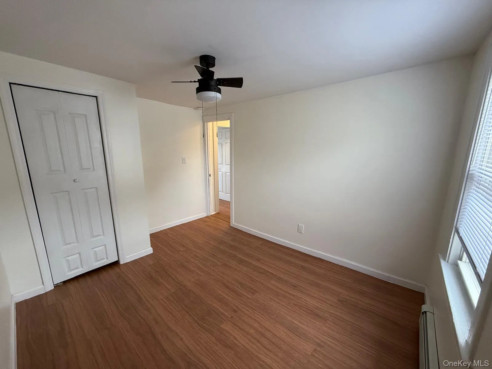 Unfurnished bedroom with ceiling fan, dark wood-type flooring, a closet, and a baseboard heating unit Unfurnished bedroom with ceiling fan, dark wood-type flooring, a closet, and a baseboard heating unit