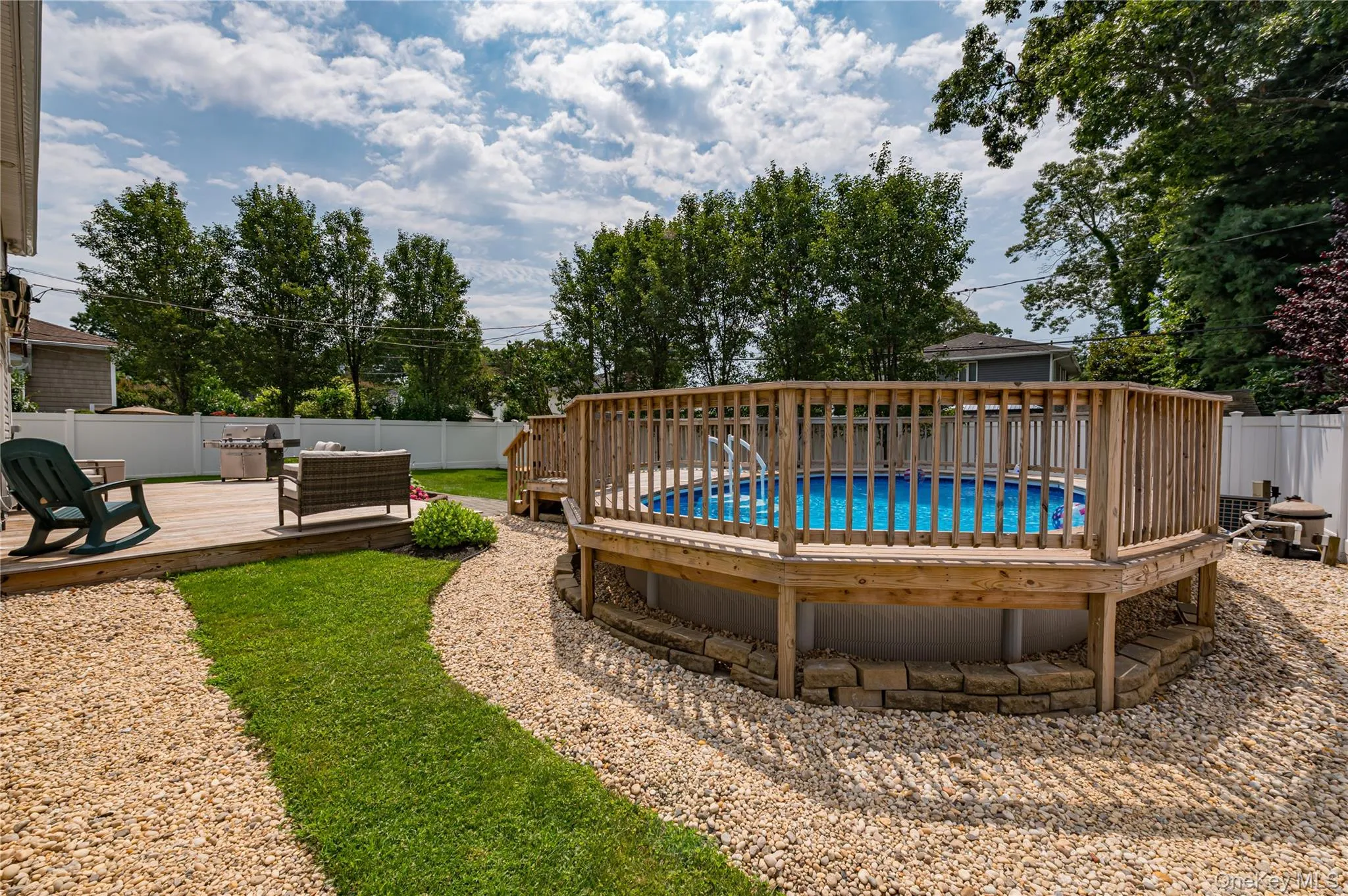 Expansive backyard featuring a built-in above ground pool with a surrounding wood deck and railing, a separate wood-deck patio, a manicured lawn path, and gravel landscaping Expansive backyard featuring a built-in above ground pool with a surrounding wood deck and railing, a separate wood-deck patio, a manicured lawn path, and gravel landscaping