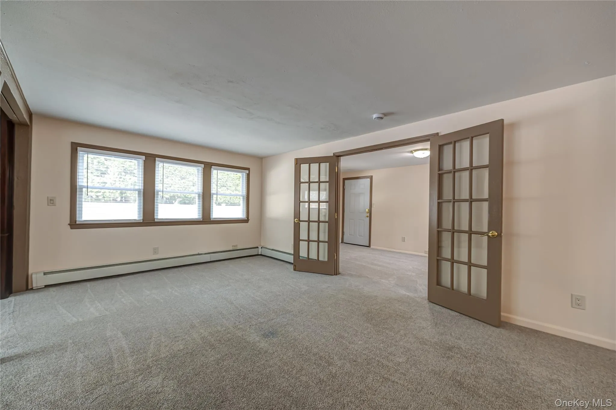 Light-colored carpeting throughout, featuring three large windows with blinds, baseboard heating, and French doors with grid-patterned glass panels Light-colored carpeting throughout, featuring three large windows with blinds, baseboard heating, and French doors with grid-patterned glass panels