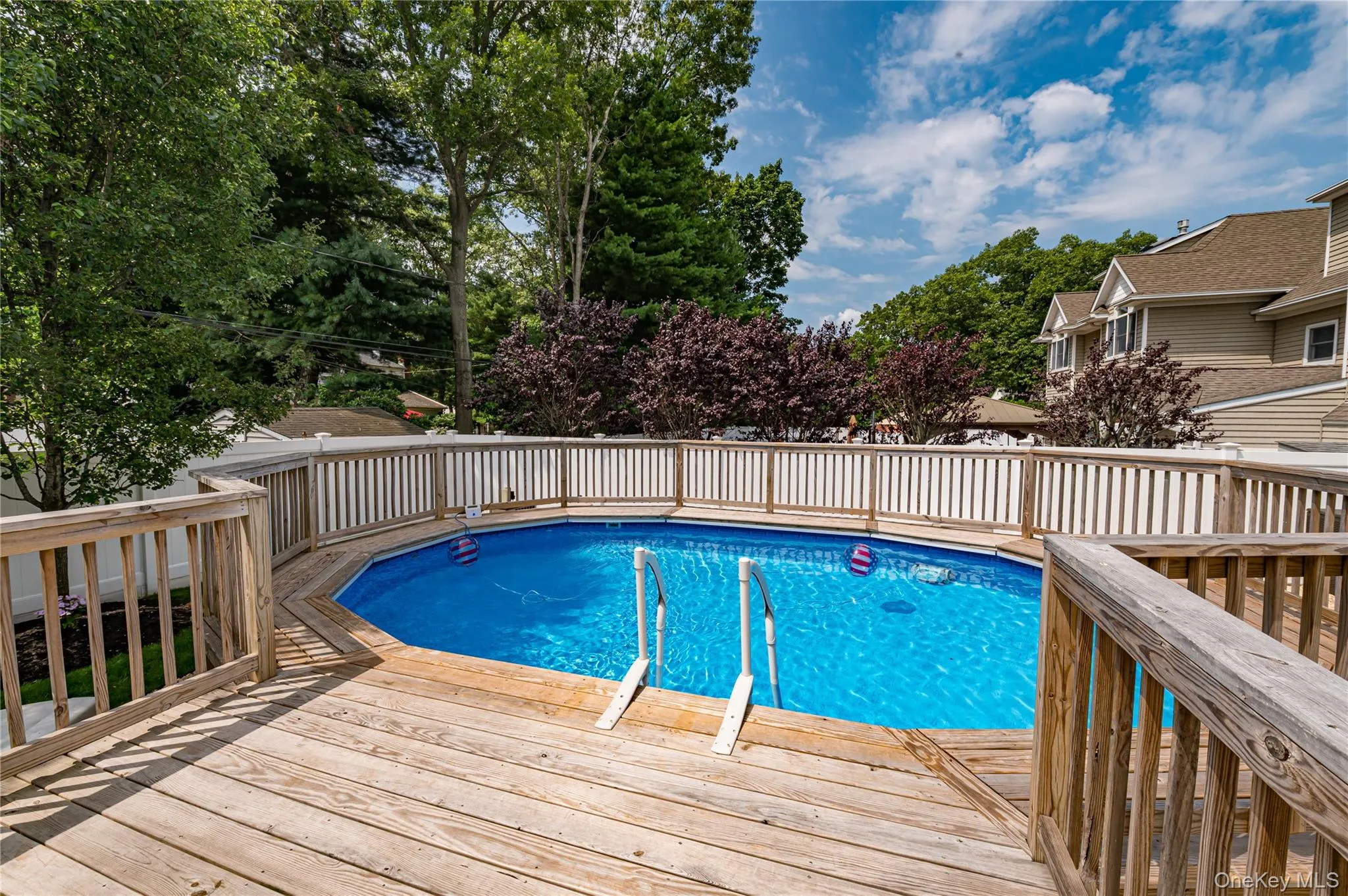 Expansive outdoor deck surrounding an above-ground pool, featuring wooden railings and a white picket fence Expansive outdoor deck surrounding an above-ground pool, featuring wooden railings and a white picket fence