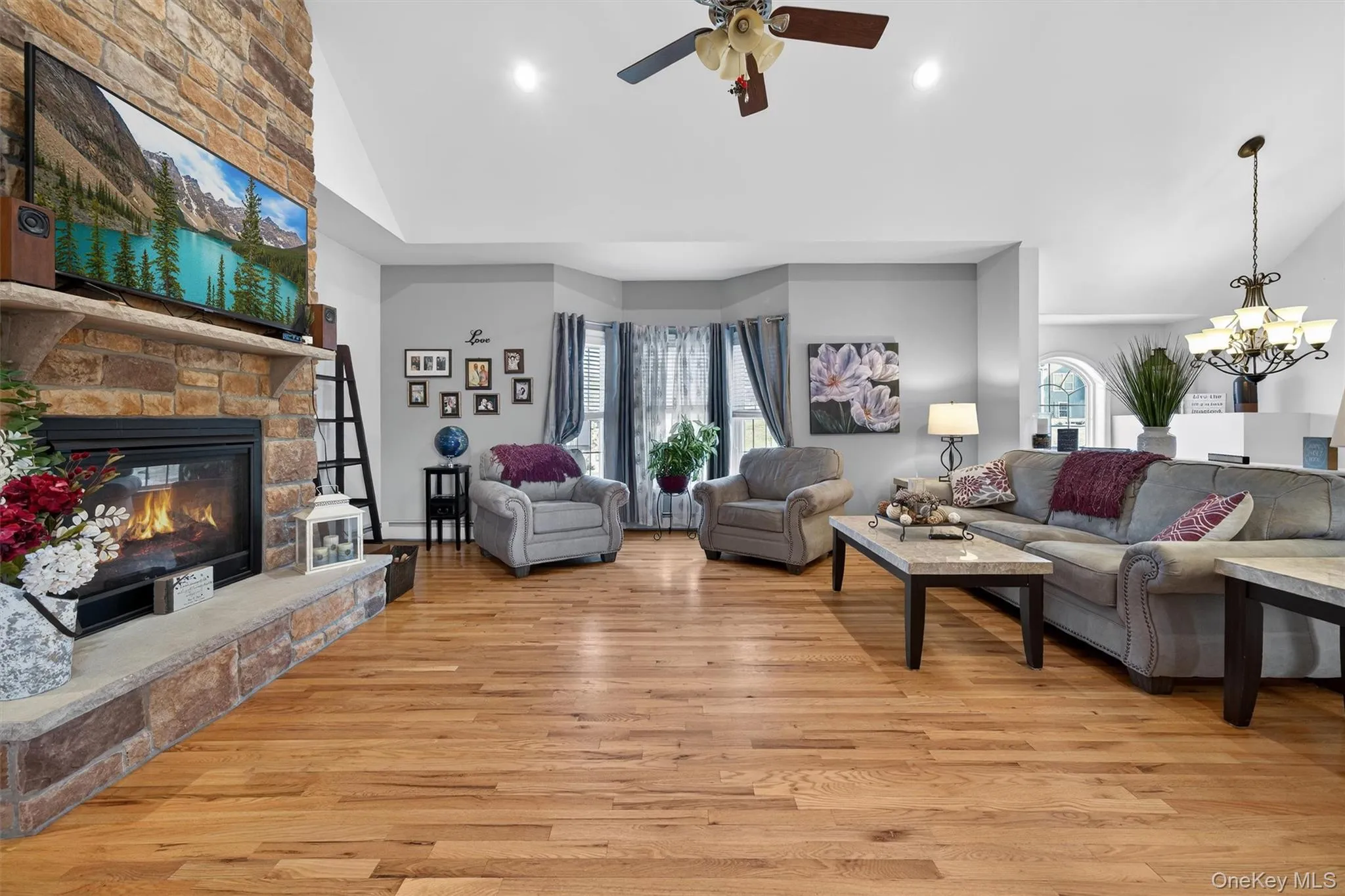 Living area featuring vaulted ceiling, a ceiling fan, a stone fireplace, light wood-type flooring, and hanging lights Living area featuring vaulted ceiling, a ceiling fan, a stone fireplace, light wood-type flooring, and hanging lights