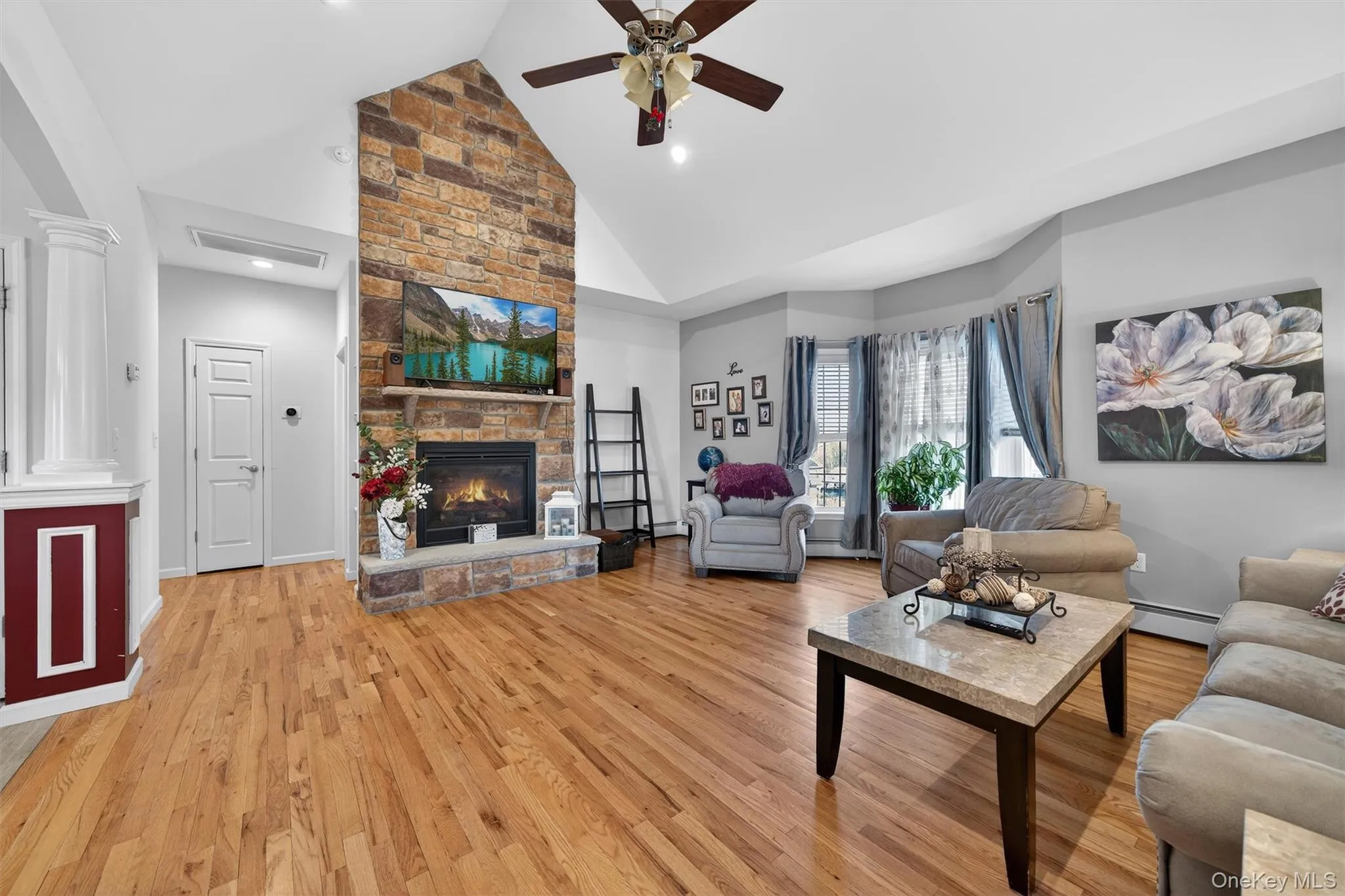 Living room featuring ceiling fan, light wood-style flooring, lofted ceiling, a stone fireplace, and a baseboard heating unit Living room featuring ceiling fan, light wood-style flooring, lofted ceiling, a stone fireplace, and a baseboard heating unit
