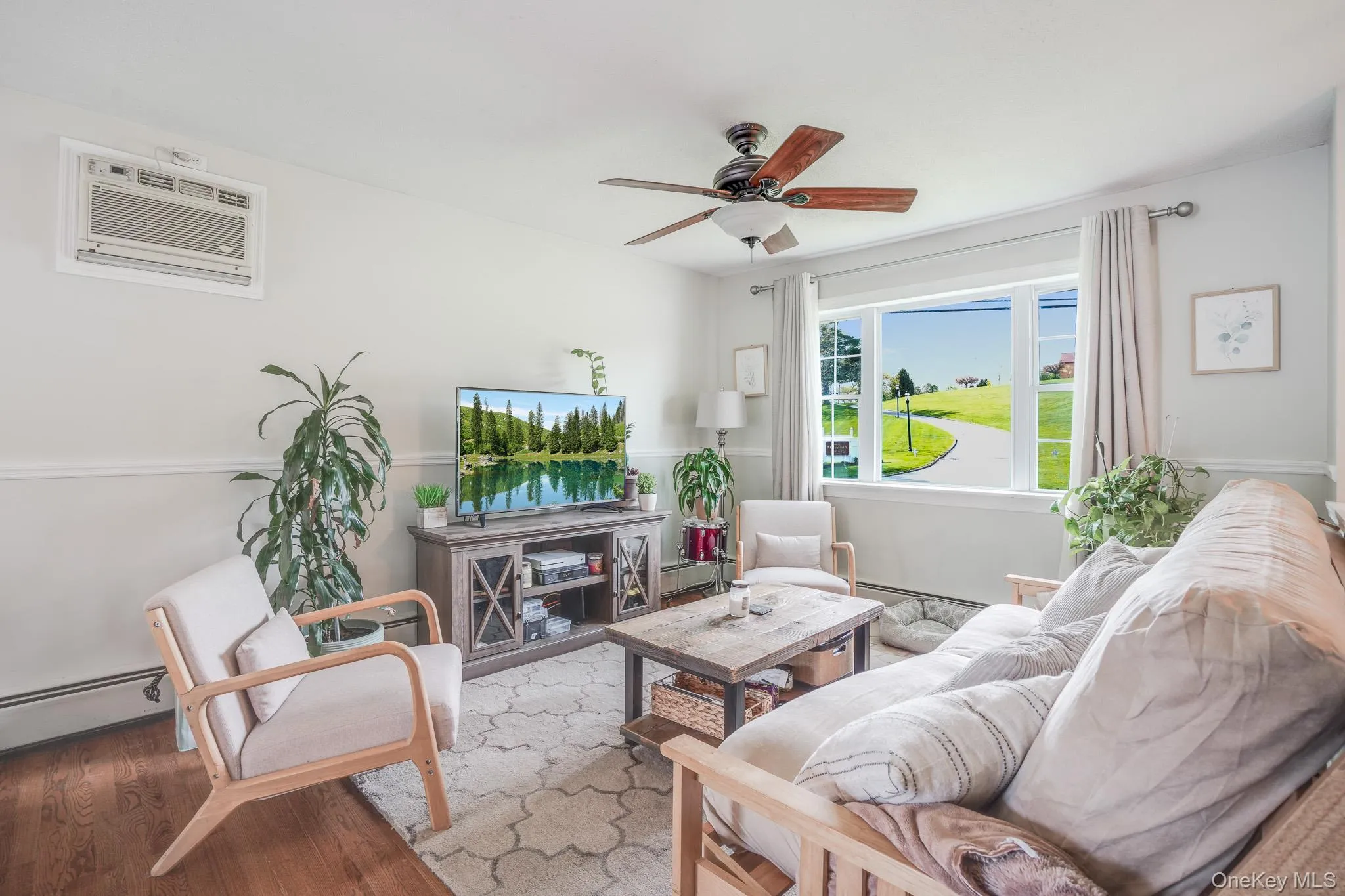 Living area featuring a large window, wood-finish flooring, a ceiling fan, and a wall-mounted air conditioning unit Living area featuring a large window, wood-finish flooring, a ceiling fan, and a wall-mounted air conditioning unit