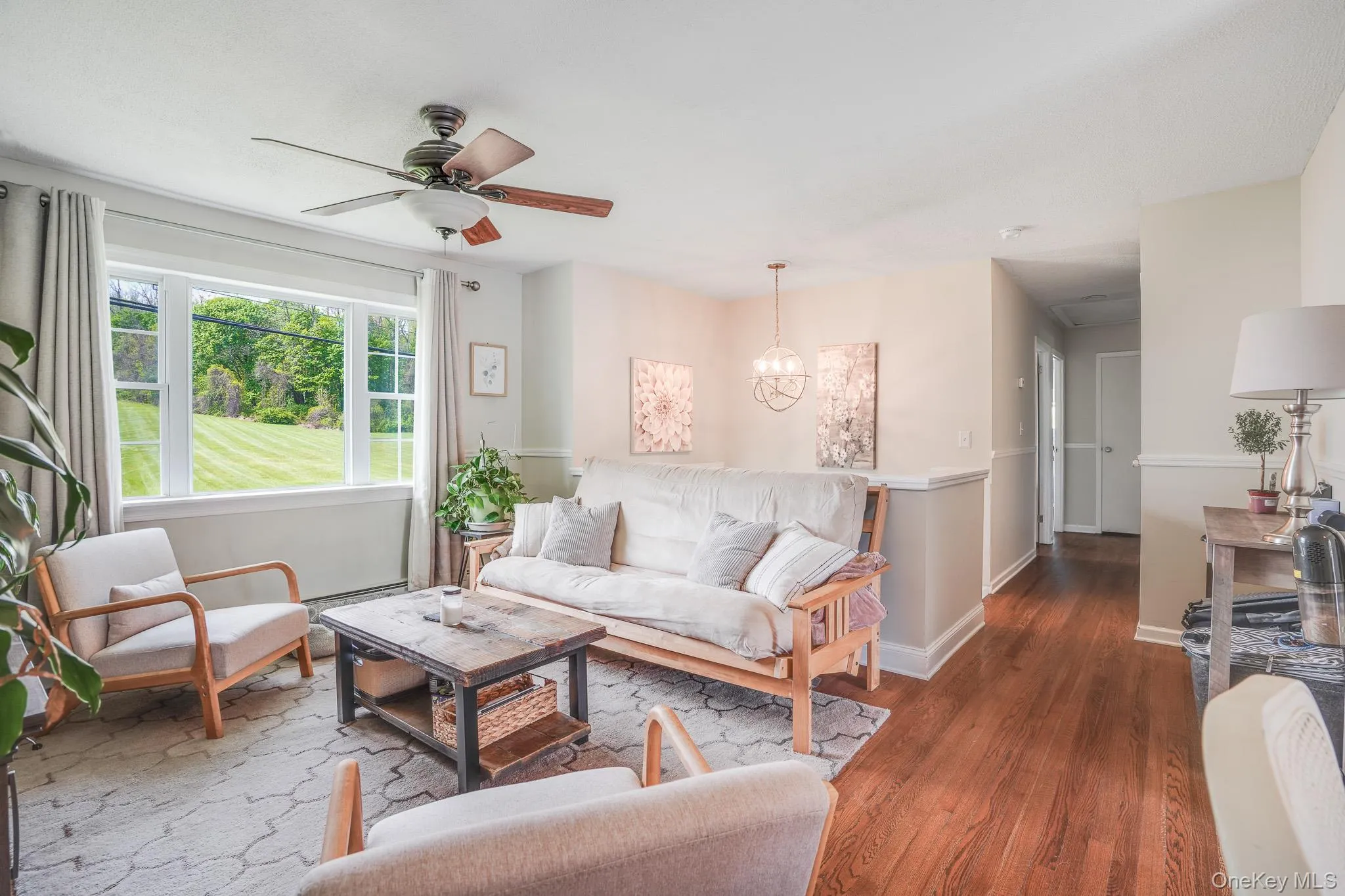 Living area featuring a large picture window overlooking a green landscape, wood-finish flooring, a ceiling fan, a contemporary chandelier, and wainscoting Living area featuring a large picture window overlooking a green landscape, wood-finish flooring, a ceiling fan, a contemporary chandelier, and wainscoting