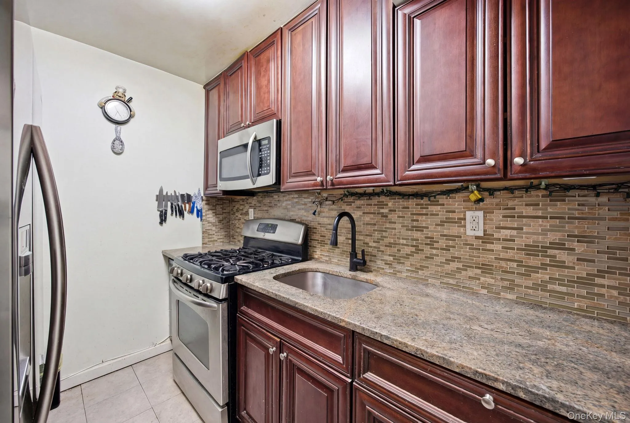Kitchen featuring light stone counters, decorative backsplash, light tile patterned floors, and reddish brown cabinets. Kitchen featuring light stone counters, decorative backsplash, light tile patterned floors, and reddish brown cabinets.