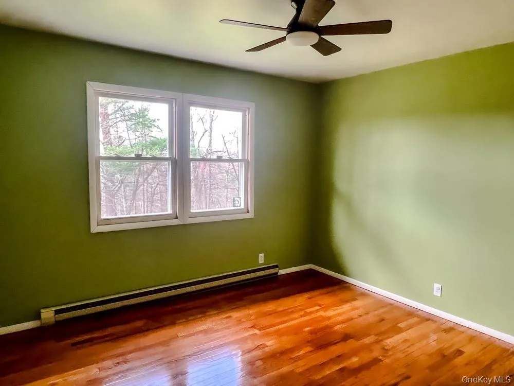 The room features polished hardwood flooring, two large windows providing views of trees, and a ceiling fan with integrated lighting The room features polished hardwood flooring, two large windows providing views of trees, and a ceiling fan with integrated lighting
