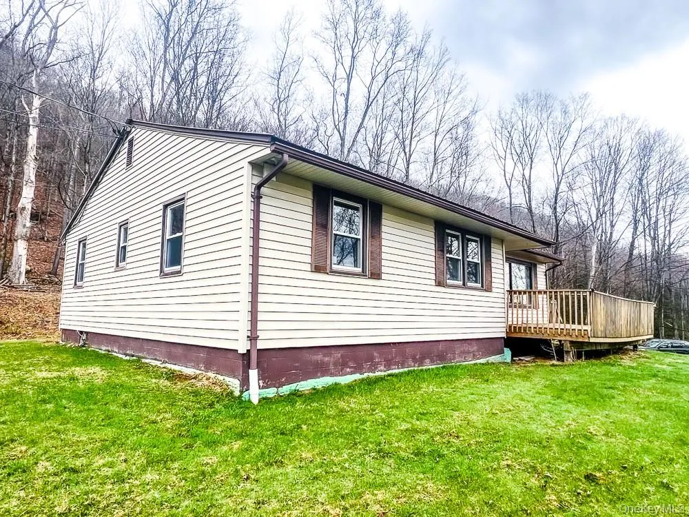 The property features a light-colored exterior with brown trim and shutters, a wooden deck, and a green lawn The property features a light-colored exterior with brown trim and shutters, a wooden deck, and a green lawn
