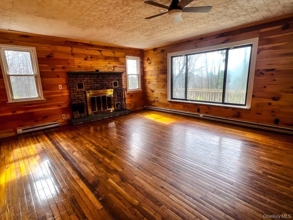 Living area featuring hardwood floors, wood paneling, a brick fireplace with a brass insert, and a ceiling fan Living area featuring hardwood floors, wood paneling, a brick fireplace with a brass insert, and a ceiling fan