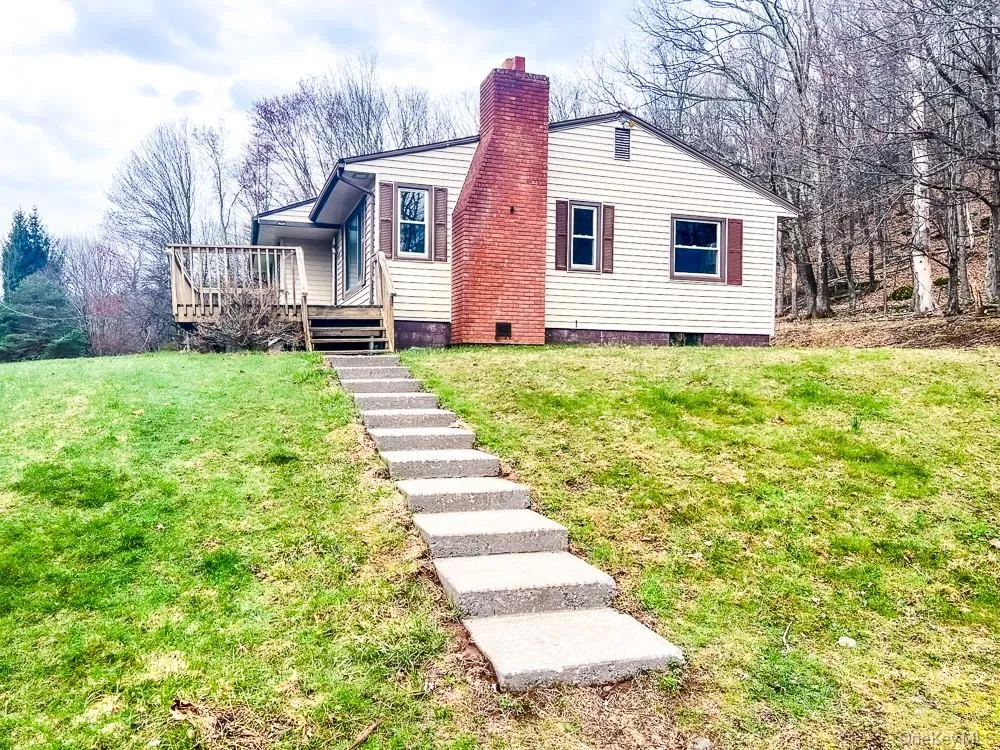 The property features a light-colored exterior with a red brick chimney, brown window shutters, and a front deck The property features a light-colored exterior with a red brick chimney, brown window shutters, and a front deck
