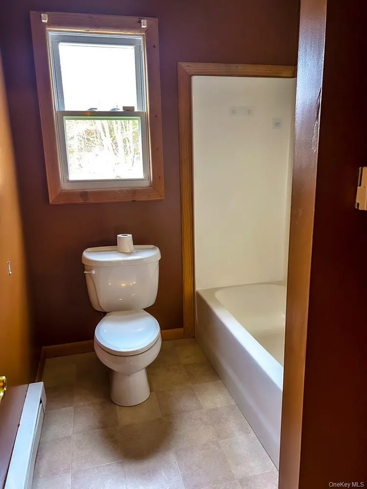 The bathroom features a toilet, a bathtub, and a window with wood trim The bathroom features a toilet, a bathtub, and a window with wood trim