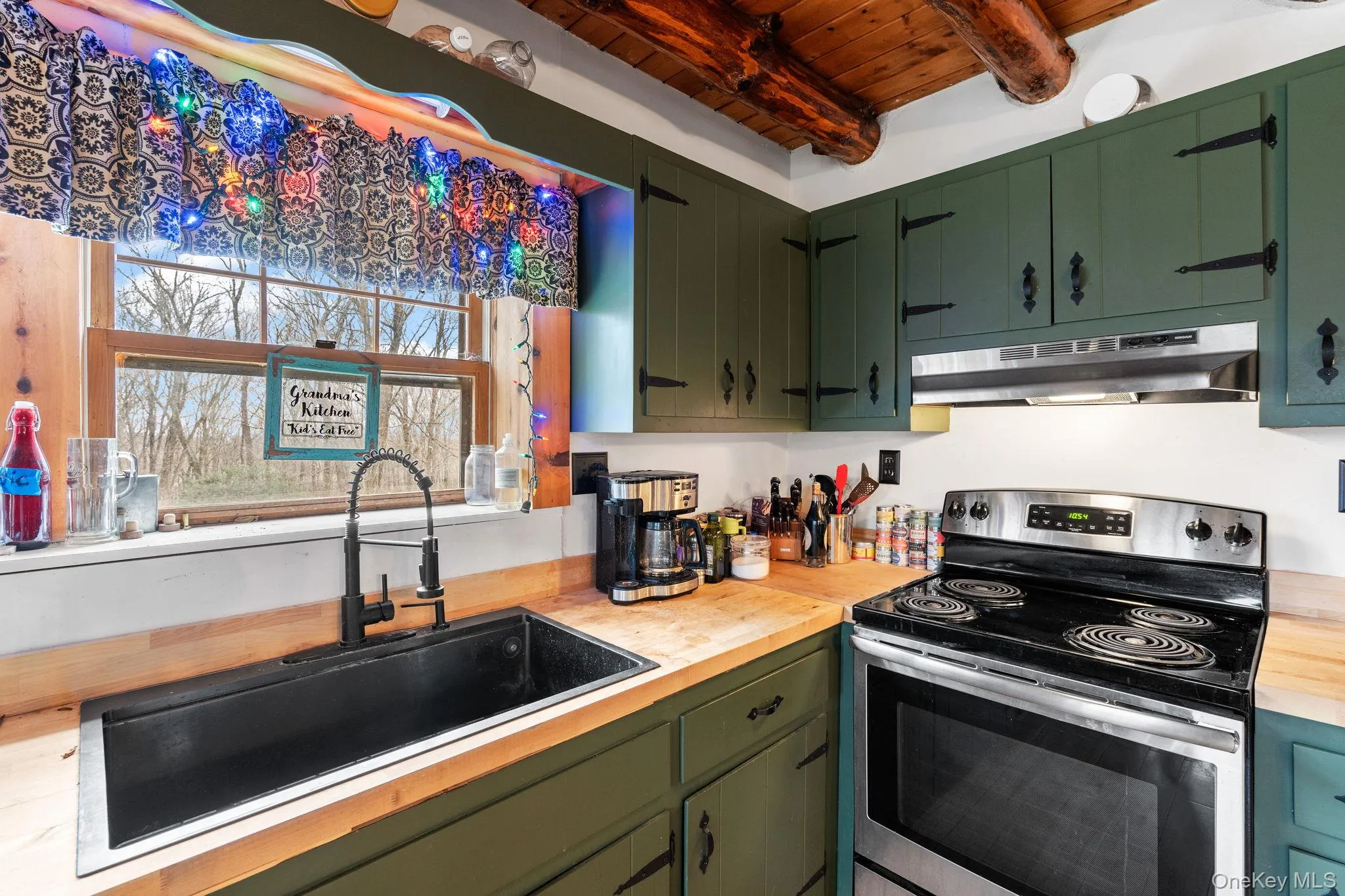 Kitchen featuring stainless steel electric range, under cabinet range hood, a wooden ceiling with exposed beams, green cabinetry, and wood counters Kitchen featuring stainless steel electric range, under cabinet range hood, a wooden ceiling with exposed beams, green cabinetry, and wood counters