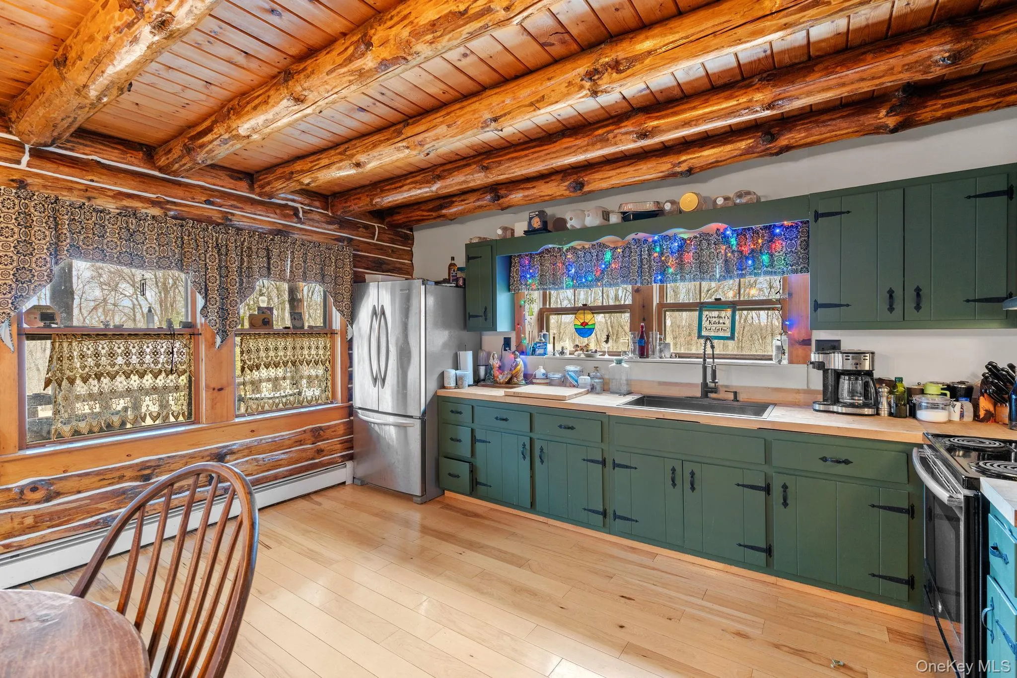 Kitchen featuring stainless steel appliances, green cabinetry, a wood ceiling with exposed beams, log walls, and a baseboard radiator Kitchen featuring stainless steel appliances, green cabinetry, a wood ceiling with exposed beams, log walls, and a baseboard radiator