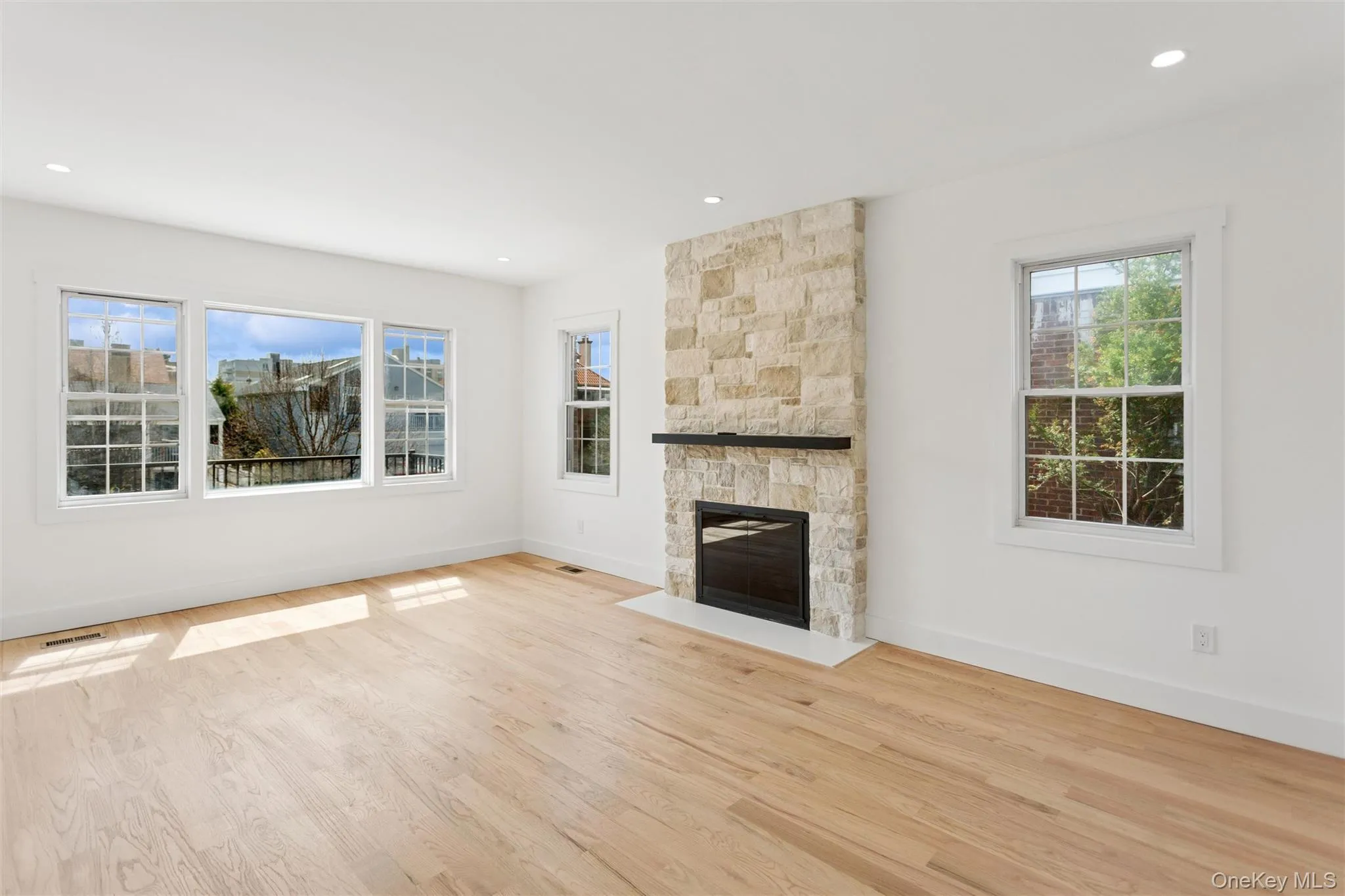 This bright living space features light wood flooring, a stone fireplace with a dark mantel, and multiple windows providing natural light This bright living space features light wood flooring, a stone fireplace with a dark mantel, and multiple windows providing natural light