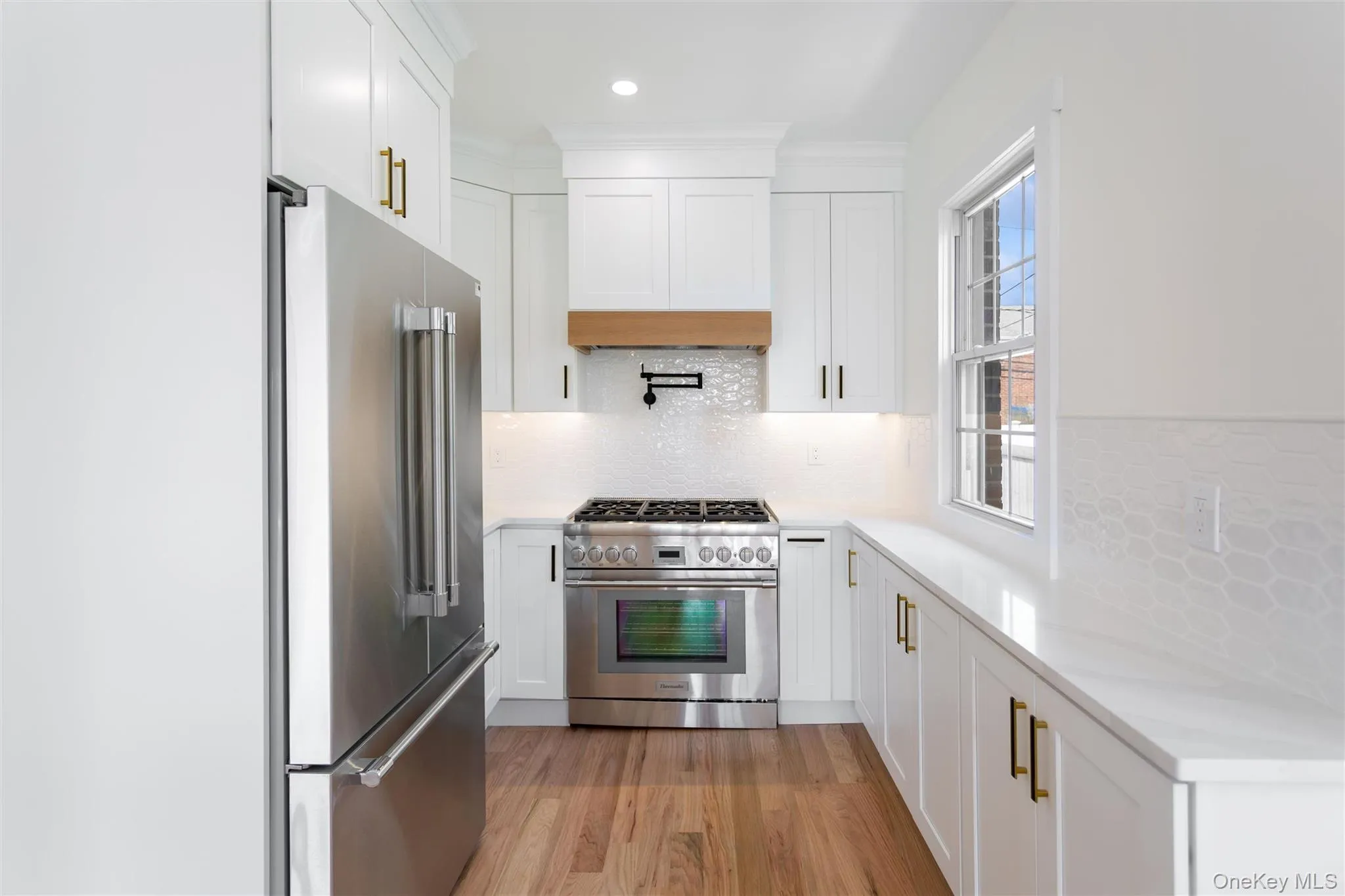 The kitchen features white cabinetry with gold-toned hardware, stainless steel appliances, light-colored countertops, and hardwood flooring The kitchen features white cabinetry with gold-toned hardware, stainless steel appliances, light-colored countertops, and hardwood flooring