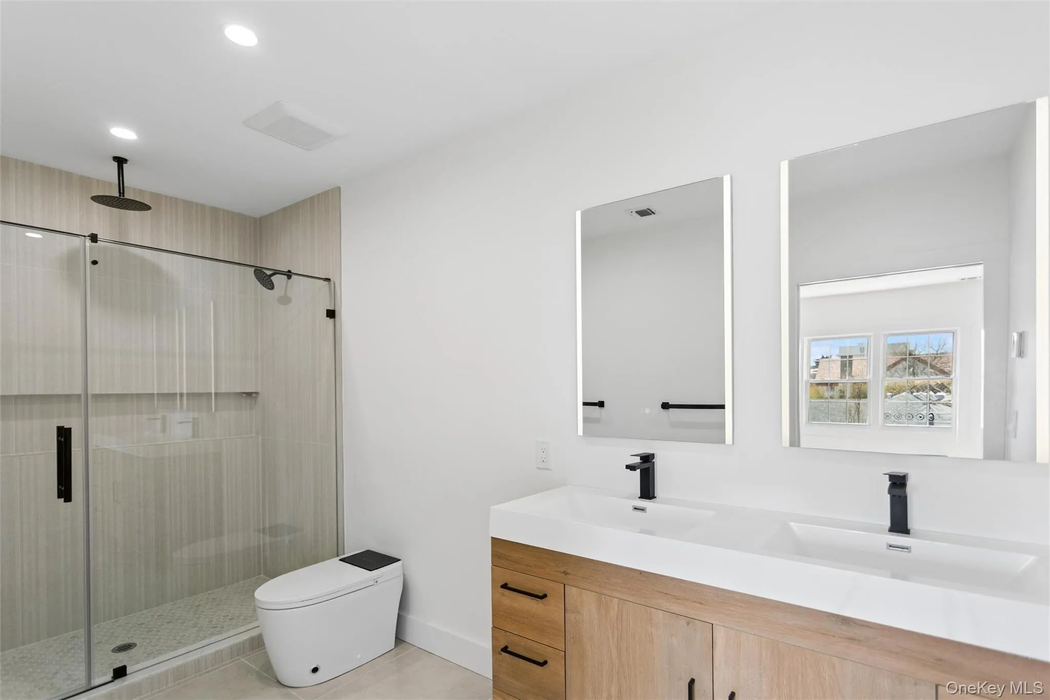 This bathroom features a spacious glass-enclosed shower with a rain showerhead, a dual vanity with rectangular sinks and dark fixtures, and illuminated mirrors This bathroom features a spacious glass-enclosed shower with a rain showerhead, a dual vanity with rectangular sinks and dark fixtures, and illuminated mirrors