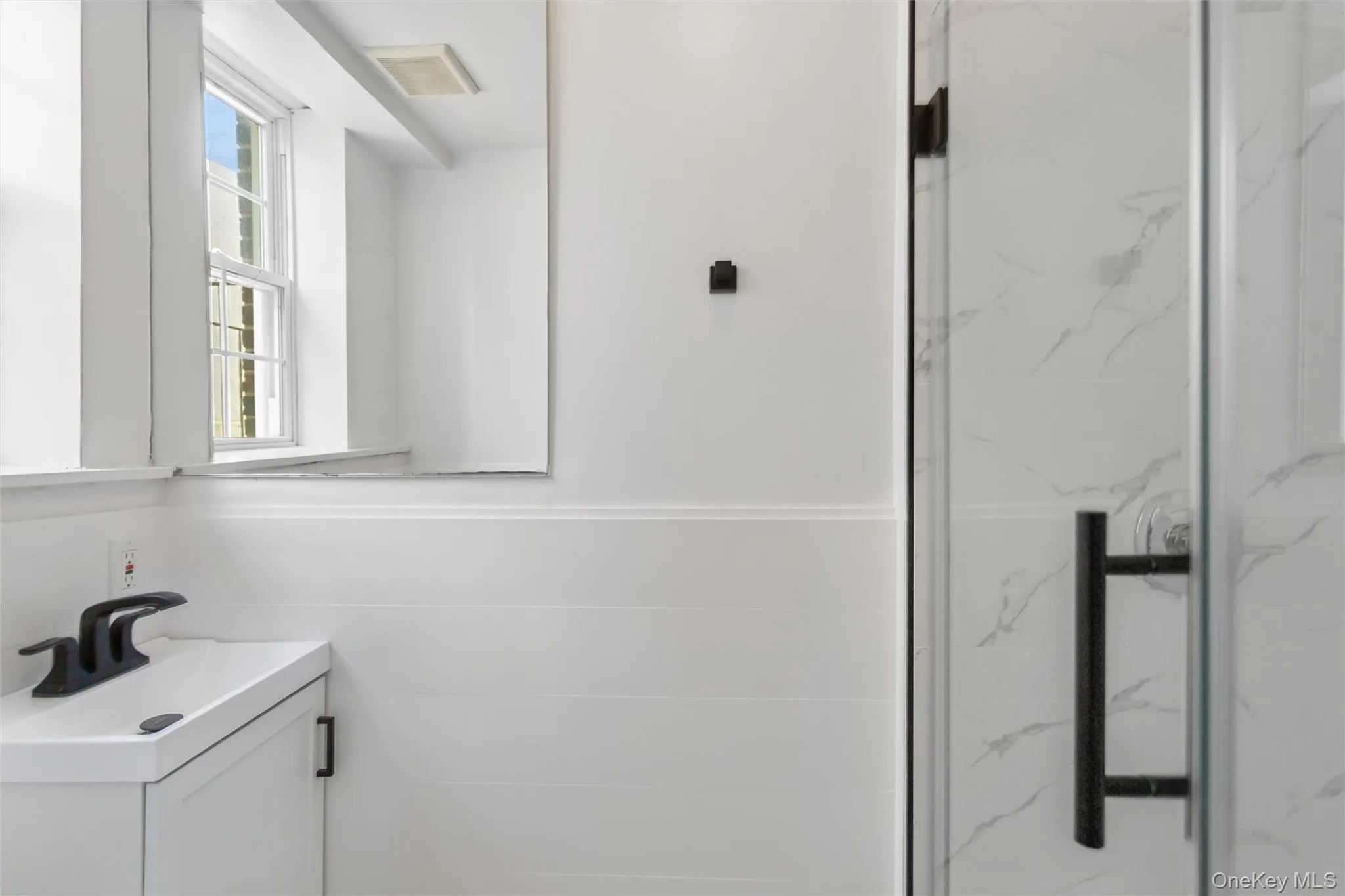 The bathroom features a modern vanity with a sink and a dark-toned faucet, a mirror, and a shower with a glass door and dark hardware The bathroom features a modern vanity with a sink and a dark-toned faucet, a mirror, and a shower with a glass door and dark hardware
