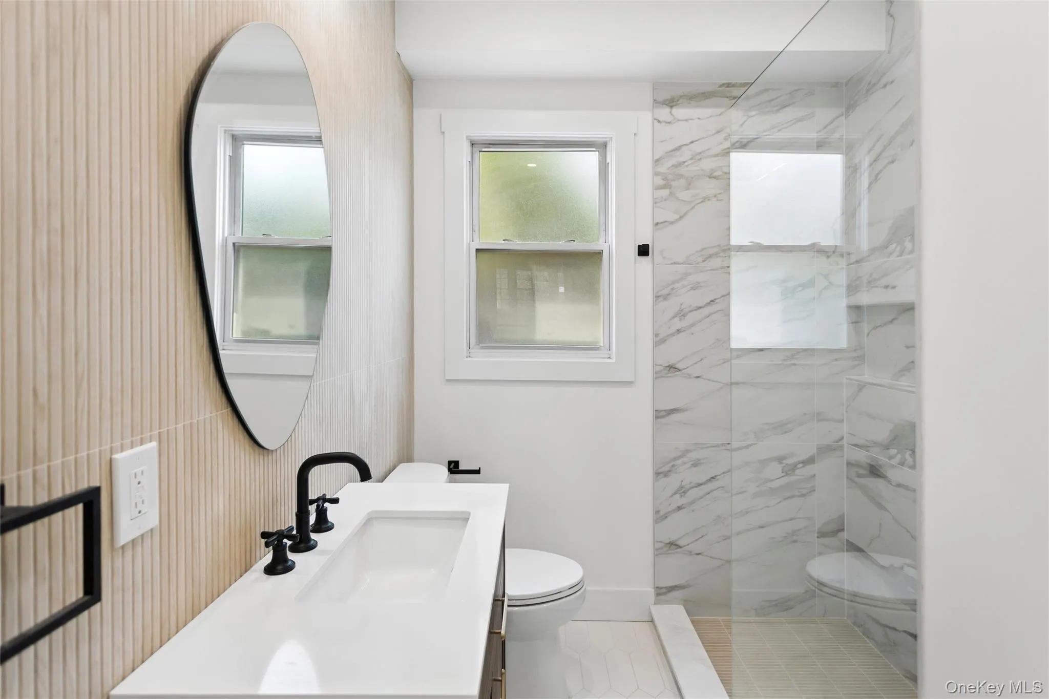 Bathroom featuring a sleek white vanity with a black faucet, a contemporary oval mirror, and a spacious shower with marble-look tiled walls and a clear glass enclosure Bathroom featuring a sleek white vanity with a black faucet, a contemporary oval mirror, and a spacious shower with marble-look tiled walls and a clear glass enclosure