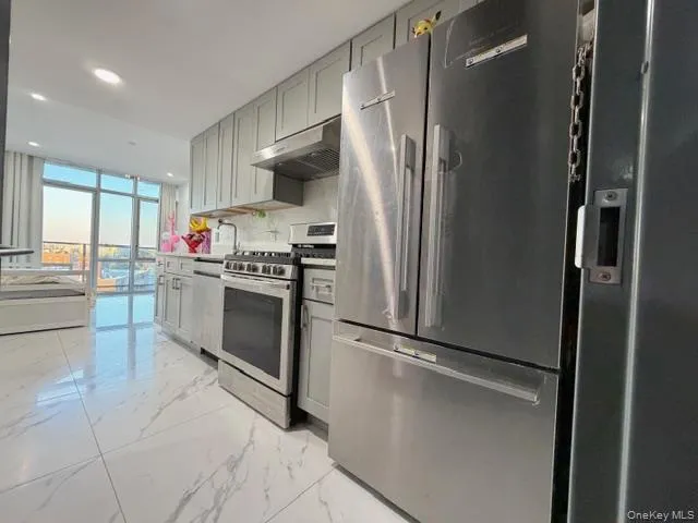 Kitchen featuring appliances with stainless steel finishes, light marble finish floors, floor to ceiling windows, gray cabinetry, and under cabinet range hood Kitchen featuring appliances with stainless steel finishes, light marble finish floors, floor to ceiling windows, gray cabinetry, and under cabinet range hood