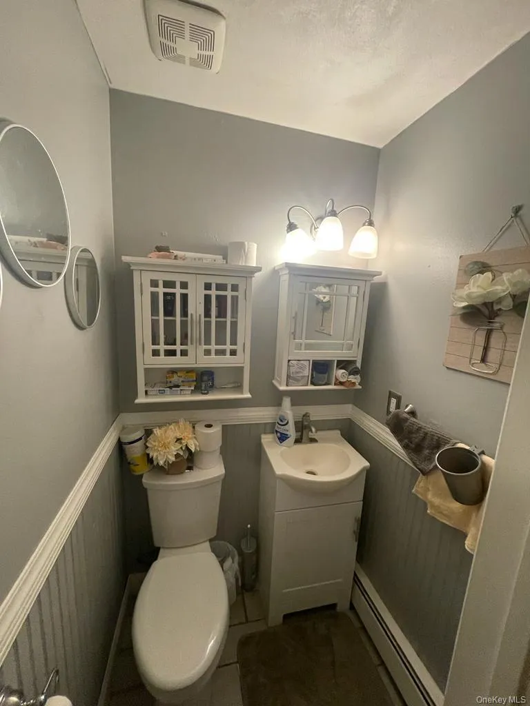 Half bath featuring visible vents, a baseboard heating unit, toilet, wainscoting, and vanity Half bath featuring visible vents, a baseboard heating unit, toilet, wainscoting, and vanity