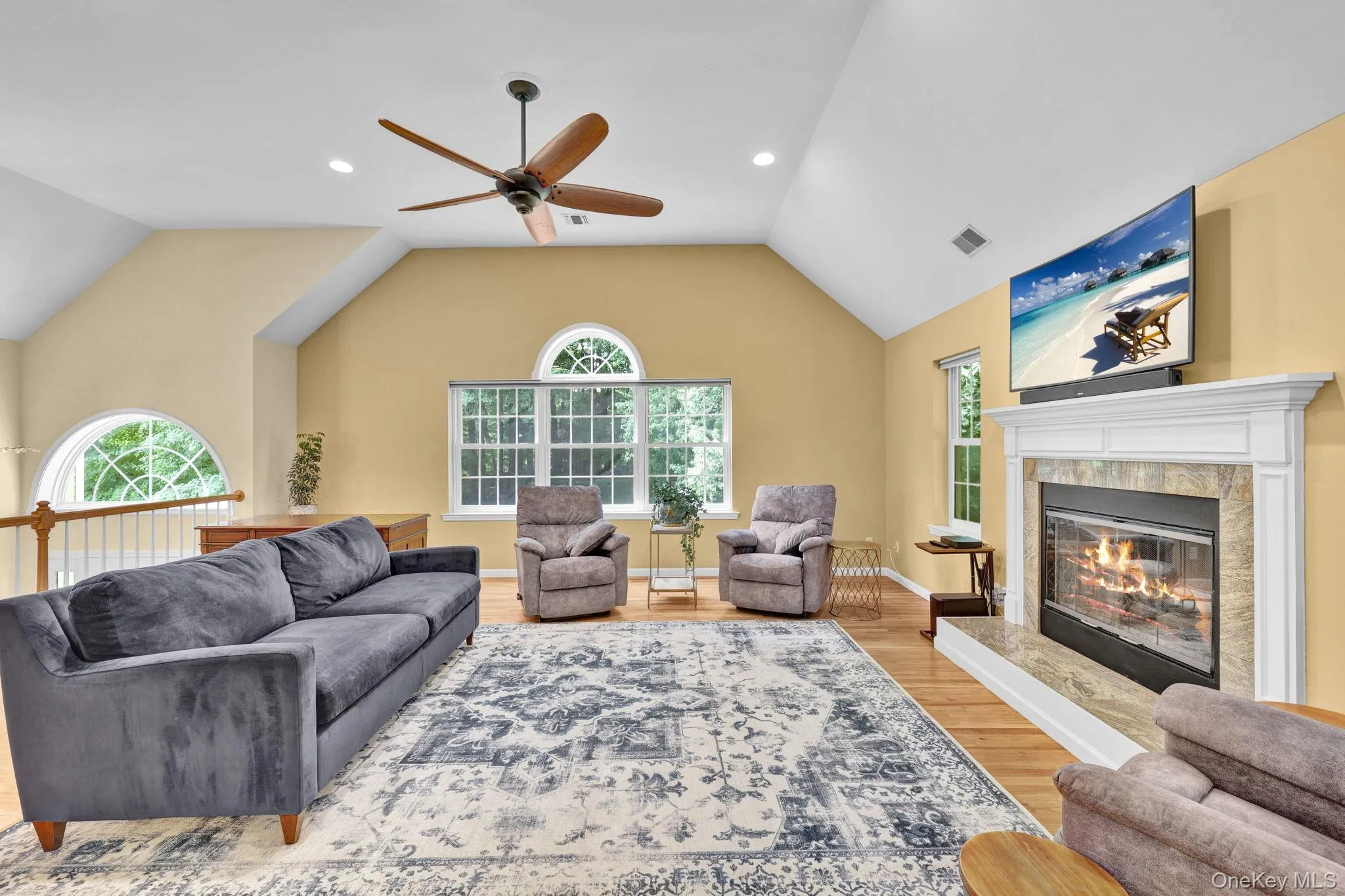 Open-concept living area featuring a vaulted ceiling with a ceiling fan, a fireplace with a stone surround and white mantel, hardwood flooring, and multiple windows including arched transoms Open-concept living area featuring a vaulted ceiling with a ceiling fan, a fireplace with a stone surround and white mantel, hardwood flooring, and multiple windows including arched transoms