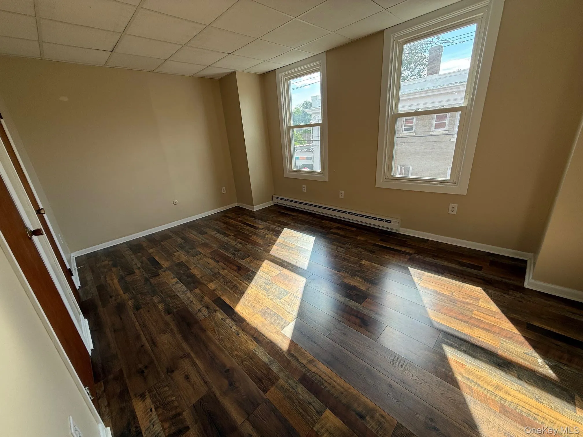 Empty room featuring dark wood-type flooring, a baseboard radiator, and a drop ceiling Empty room featuring dark wood-type flooring, a baseboard radiator, and a drop ceiling