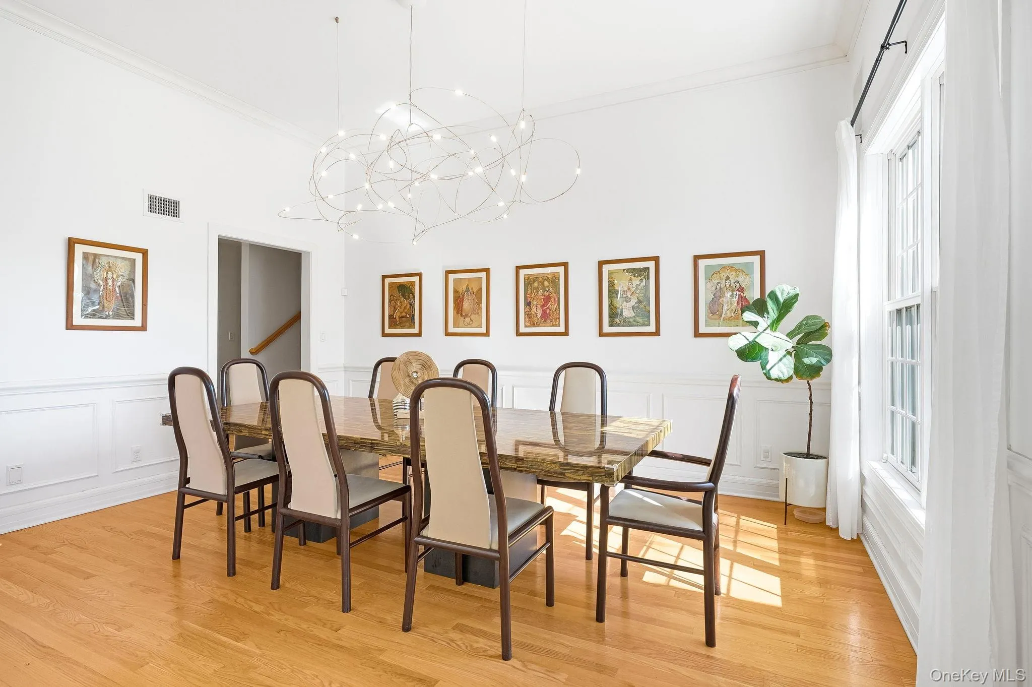The dining room features gorgeous hardwood flooring, white wainscoting, an oversized window, a modern chandelier, and crown molding, and connects directly to the kitchen. The dining room features gorgeous hardwood flooring, white wainscoting, an oversized window, a modern chandelier, and crown molding, and connects directly to the kitchen.