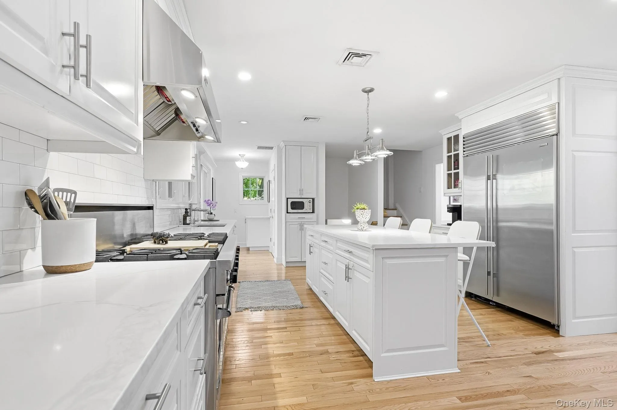 The open-concept kitchen features an expansive central island with seating, white shaker cabinetry, stainless steel appliances, and wood-finish flooring. The open-concept kitchen features an expansive central island with seating, white shaker cabinetry, stainless steel appliances, and wood-finish flooring.