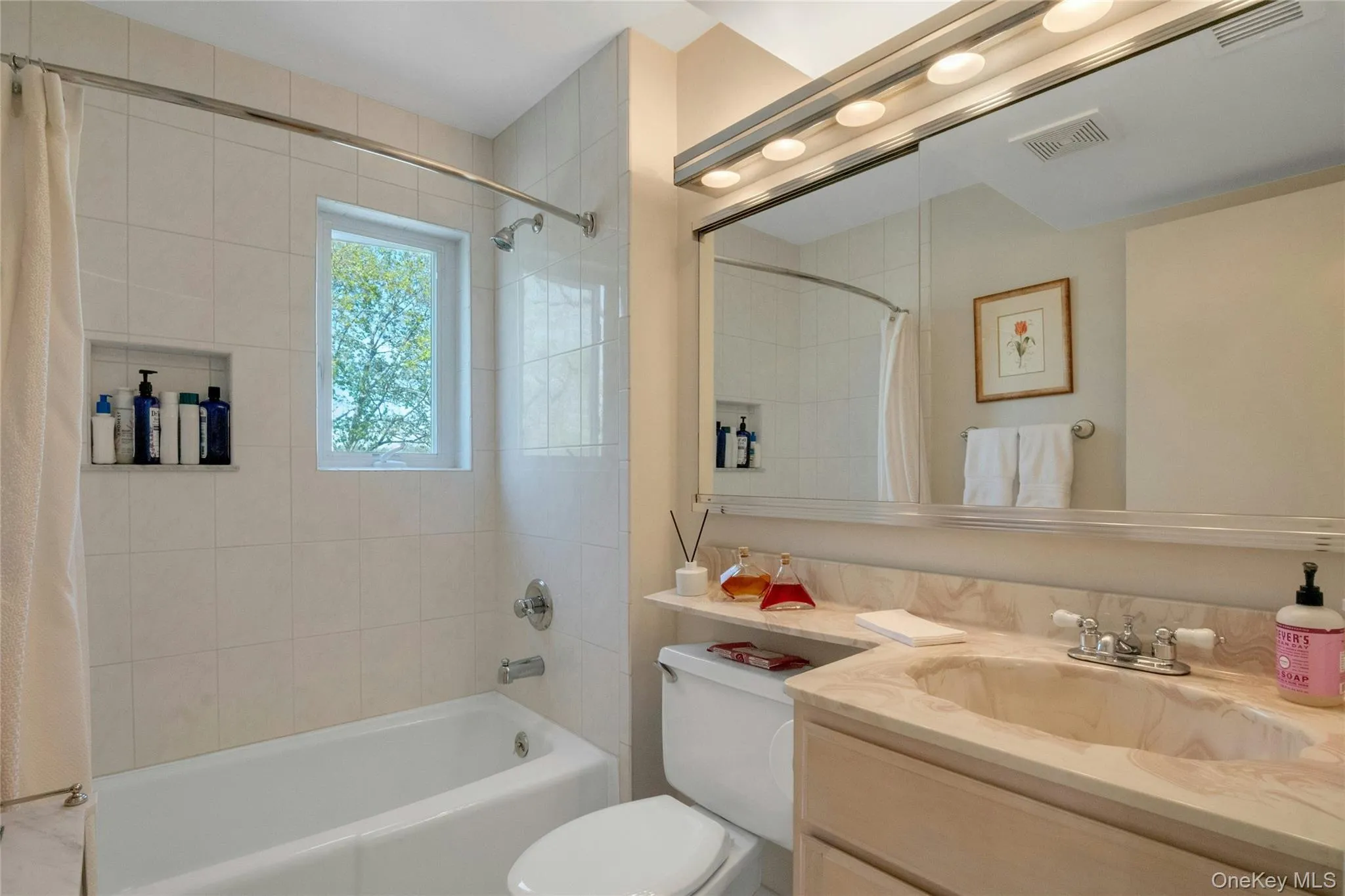 Bathroom featuring a tiled shower/tub combination with a built-in niche, a window, a vanity with a stone countertop and integrated sink, and an expansive mirror with overhead lighting Bathroom featuring a tiled shower/tub combination with a built-in niche, a window, a vanity with a stone countertop and integrated sink, and an expansive mirror with overhead lighting
