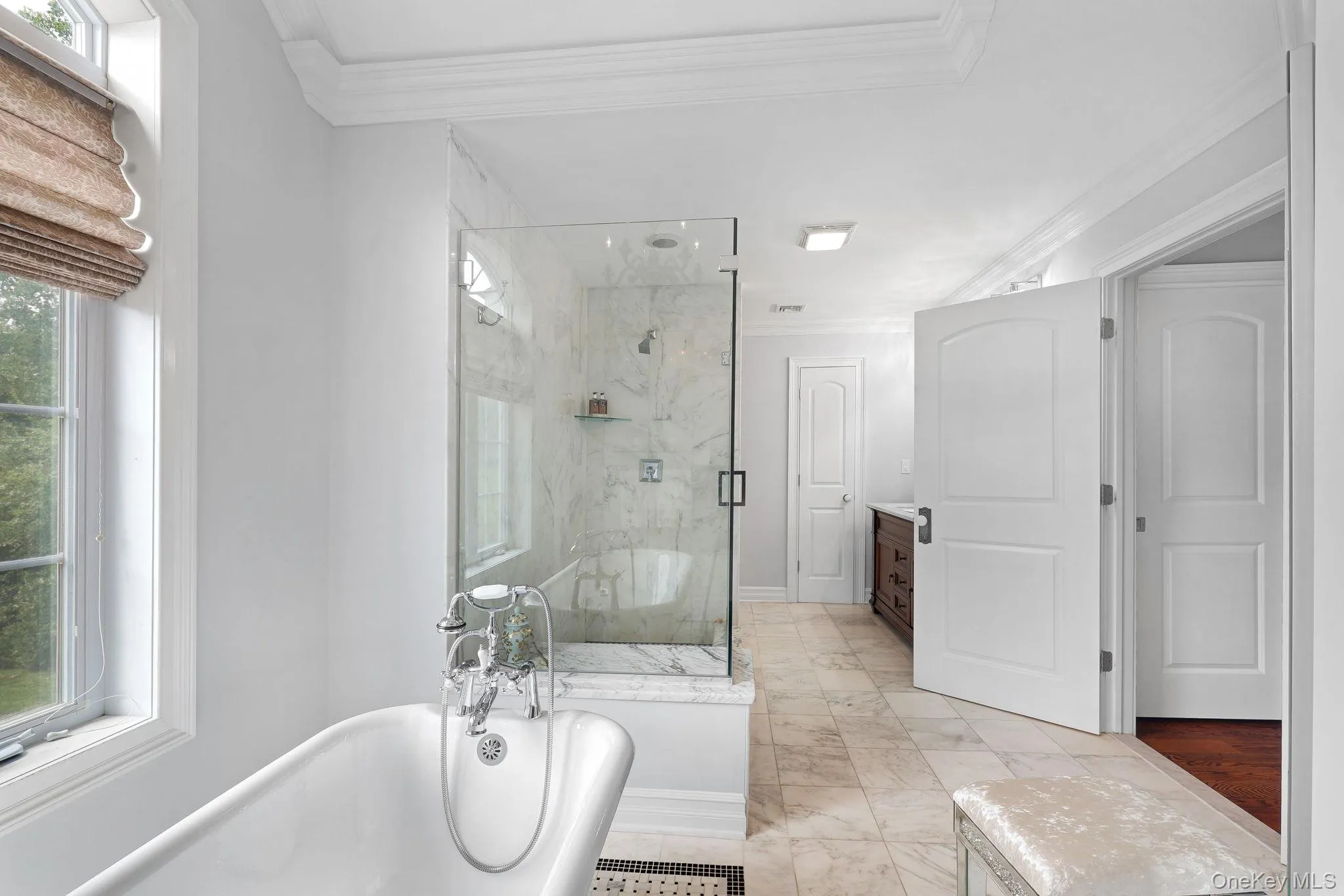 Freestanding clawfoot tub with chrome fixtures, glass-enclosed shower with marble-style tile, light-toned tile flooring, white crown molding, and a dark wood vanity Freestanding clawfoot tub with chrome fixtures, glass-enclosed shower with marble-style tile, light-toned tile flooring, white crown molding, and a dark wood vanity