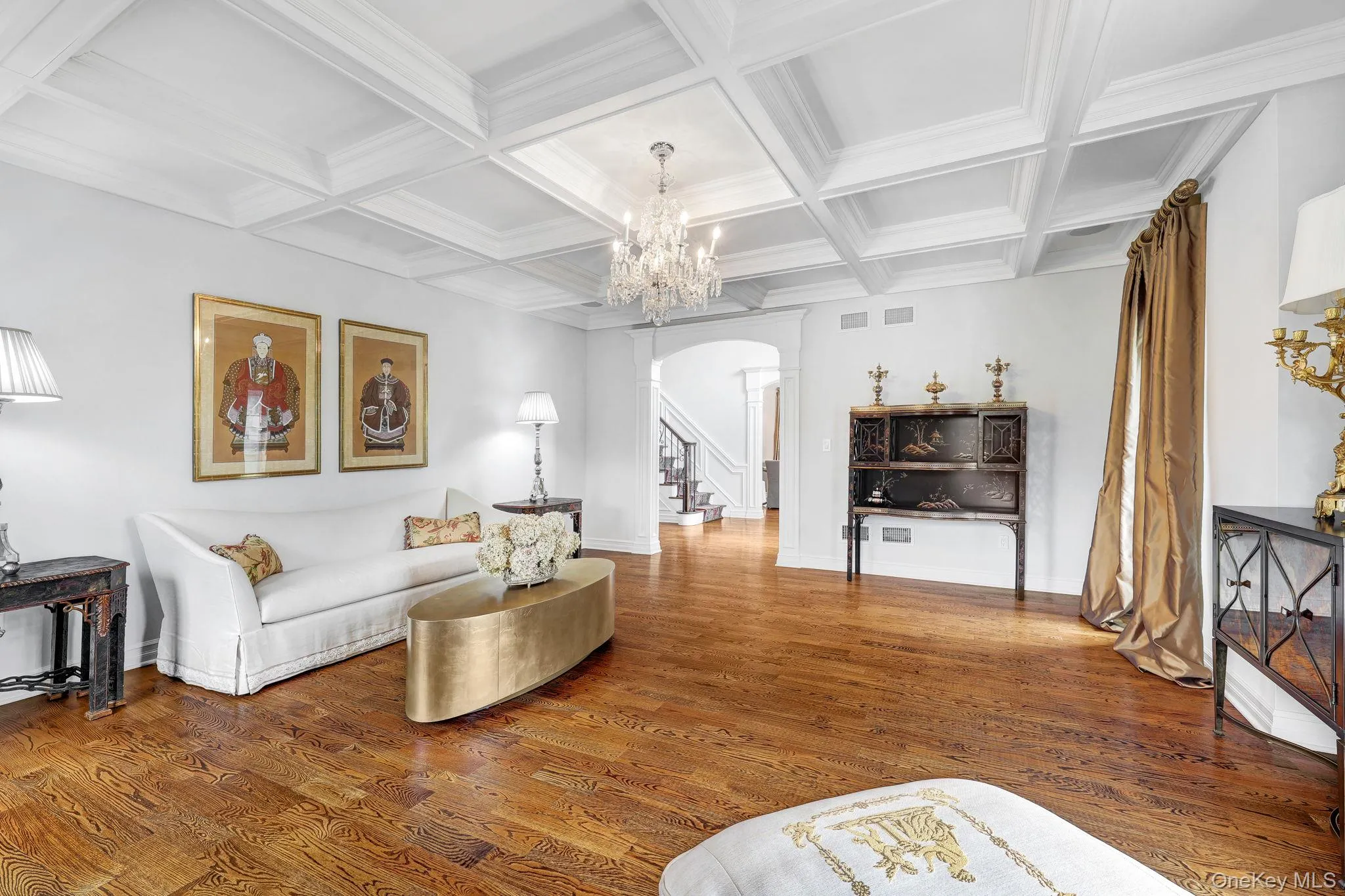 Hardwood flooring and a coffered ceiling with a crystal chandelier illuminate the living space, featuring an arched doorway and a built-in display cabinet Hardwood flooring and a coffered ceiling with a crystal chandelier illuminate the living space, featuring an arched doorway and a built-in display cabinet