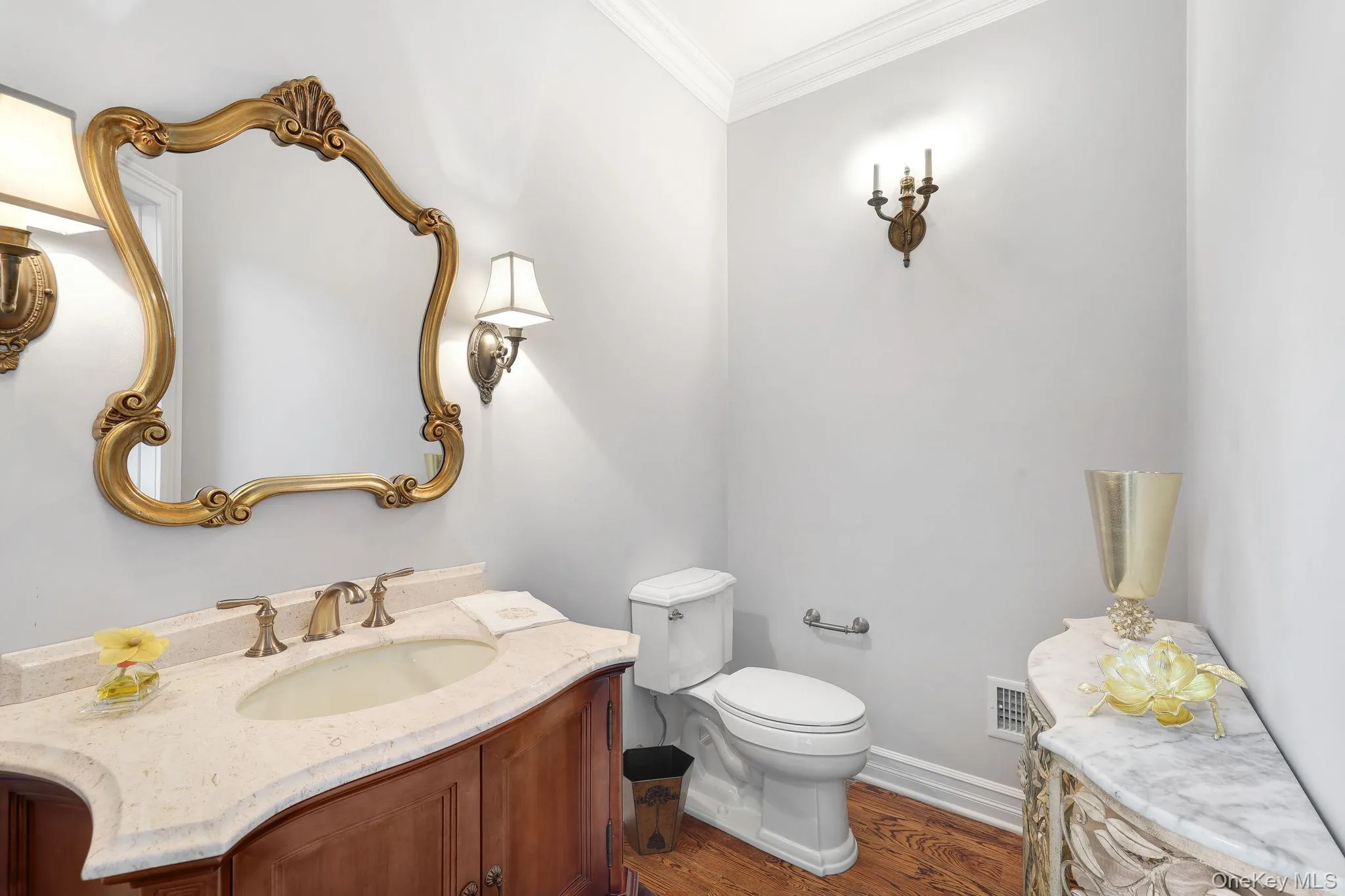First floor bathroom featuring a wood vanity with a light-toned countertop, an ornate gold-framed mirror, and wood-finish flooring First floor bathroom featuring a wood vanity with a light-toned countertop, an ornate gold-framed mirror, and wood-finish flooring