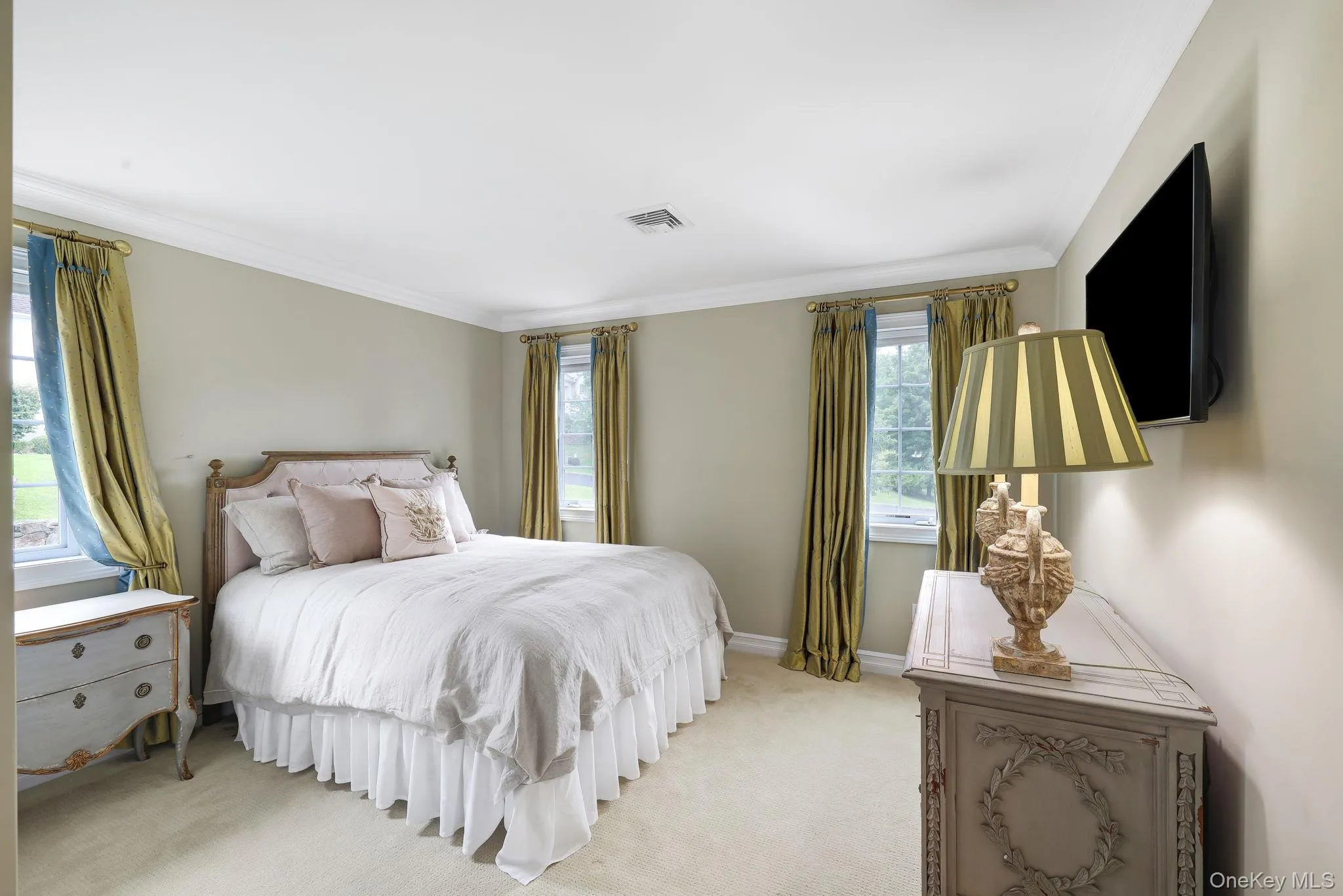 Elegant primary bedroom featuring multiple windows with natural light, neutral wall finishes, and wall-mounted flat-screen television Elegant primary bedroom featuring multiple windows with natural light, neutral wall finishes, and wall-mounted flat-screen television