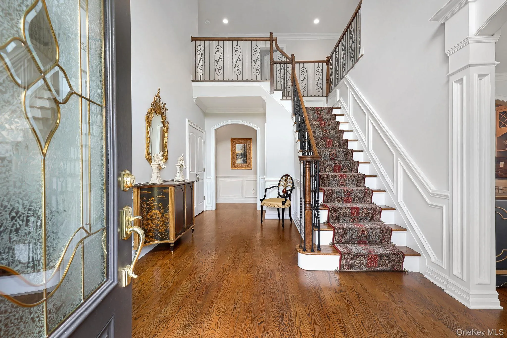 Two-story foyer with hardwood flooring, a grand staircase featuring wood and wrought iron railings, decorative wainscoting, and an arched entryway Two-story foyer with hardwood flooring, a grand staircase featuring wood and wrought iron railings, decorative wainscoting, and an arched entryway