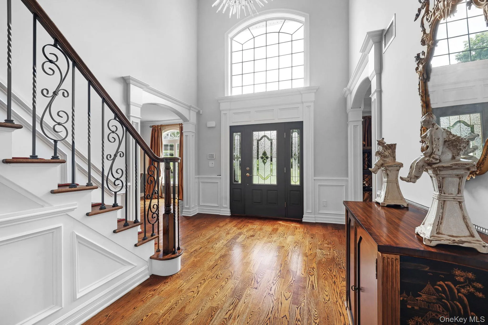 Two-story entryway with a decorative arched window, dark wood-finish flooring, and a staircase featuring intricate wrought iron balusters Two-story entryway with a decorative arched window, dark wood-finish flooring, and a staircase featuring intricate wrought iron balusters