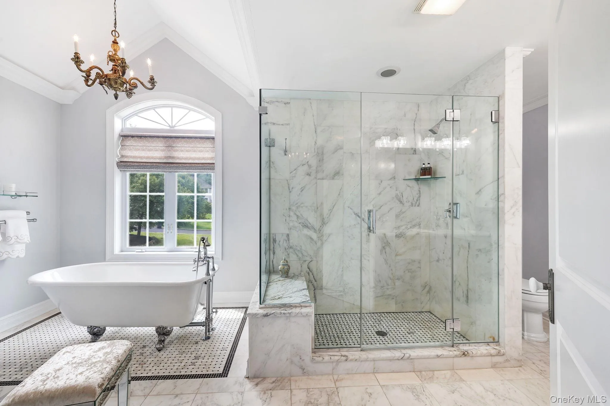 Clawfoot soaking tub, glass-enclosed shower with marble-look tile, arched window, chandelier, and a mosaic tile floor accent Clawfoot soaking tub, glass-enclosed shower with marble-look tile, arched window, chandelier, and a mosaic tile floor accent