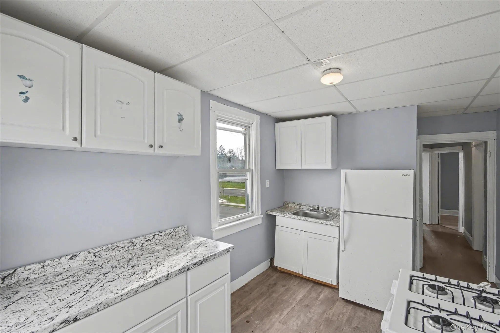 Kitchen featuring freestanding refrigerator, white cabinetry, dark wood-type flooring, and a drop ceiling Kitchen featuring freestanding refrigerator, white cabinetry, dark wood-type flooring, and a drop ceiling