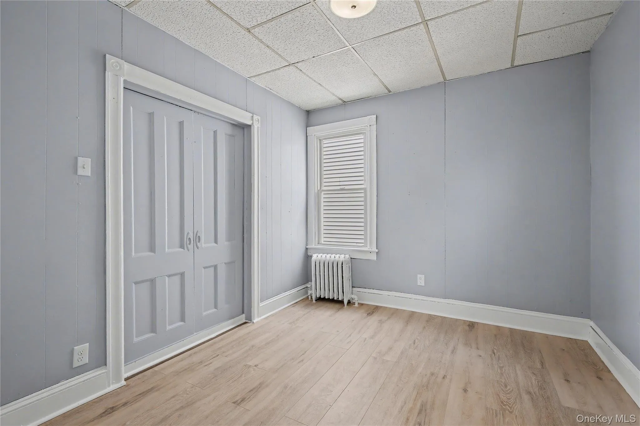 Unfurnished bedroom with a paneled ceiling, radiator heating unit, light wood-style flooring, and a closet Unfurnished bedroom with a paneled ceiling, radiator heating unit, light wood-style flooring, and a closet