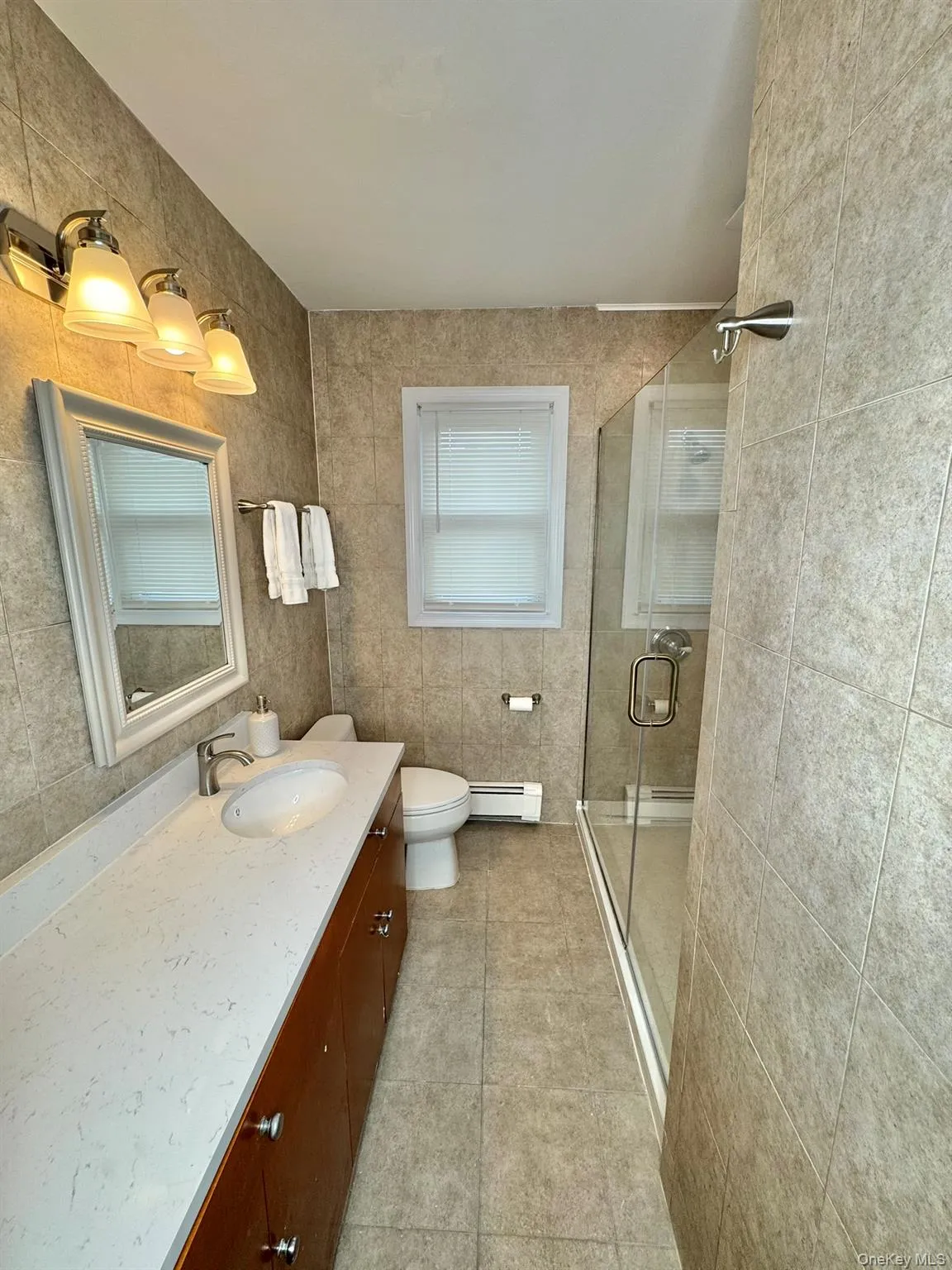 Full bath featuring vanity, a stall shower, tile walls, a baseboard radiator, and light tile patterned flooring Full bath featuring vanity, a stall shower, tile walls, a baseboard radiator, and light tile patterned flooring
