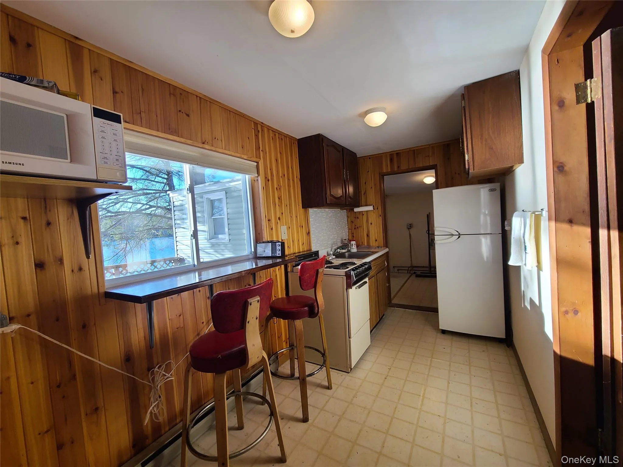 Kitchen featuring light flooring, wooden walls, white appliances, a baseboard heating unit, and a breakfast bar Kitchen featuring light flooring, wooden walls, white appliances, a baseboard heating unit, and a breakfast bar